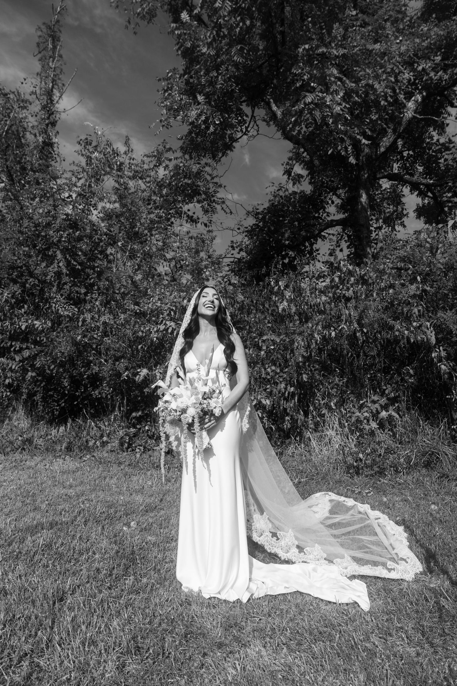 Black and white photo of a bride in a flowing gown and veil holding a bouquet, standing in a lush outdoor setting.