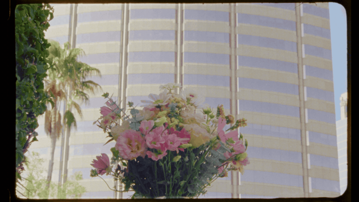 Pink and white flower bouquet in front of a striped building facade with palm trees on a sunny day.