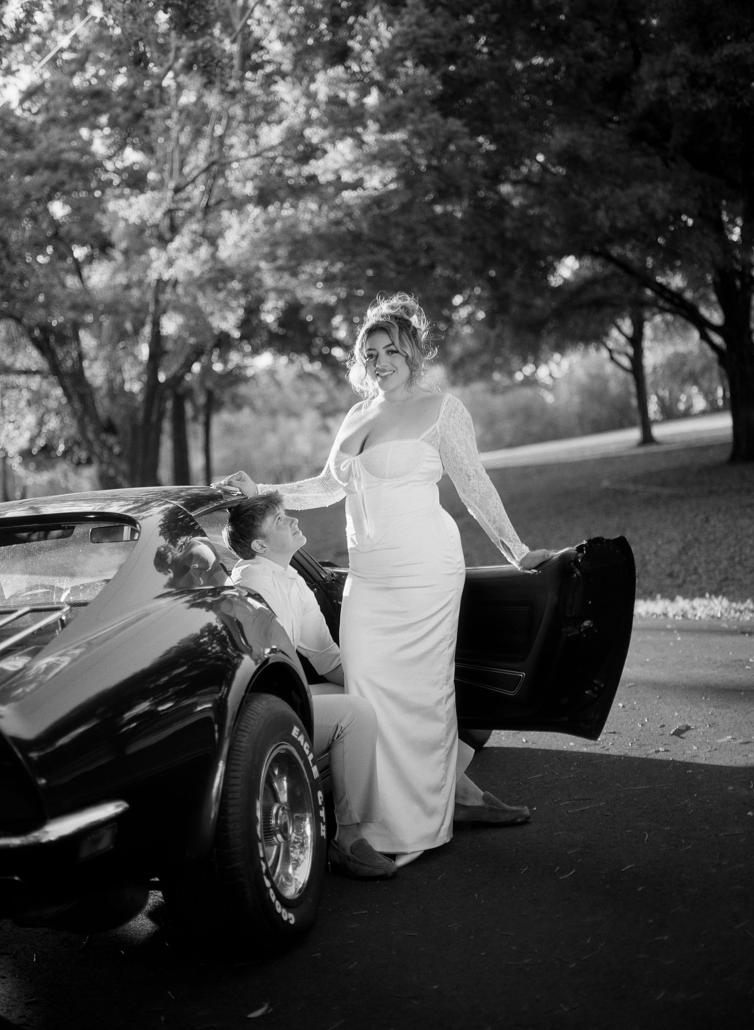 Black and white photo of a bride in a white gown standing beside a classic convertible car with a groom seated inside.