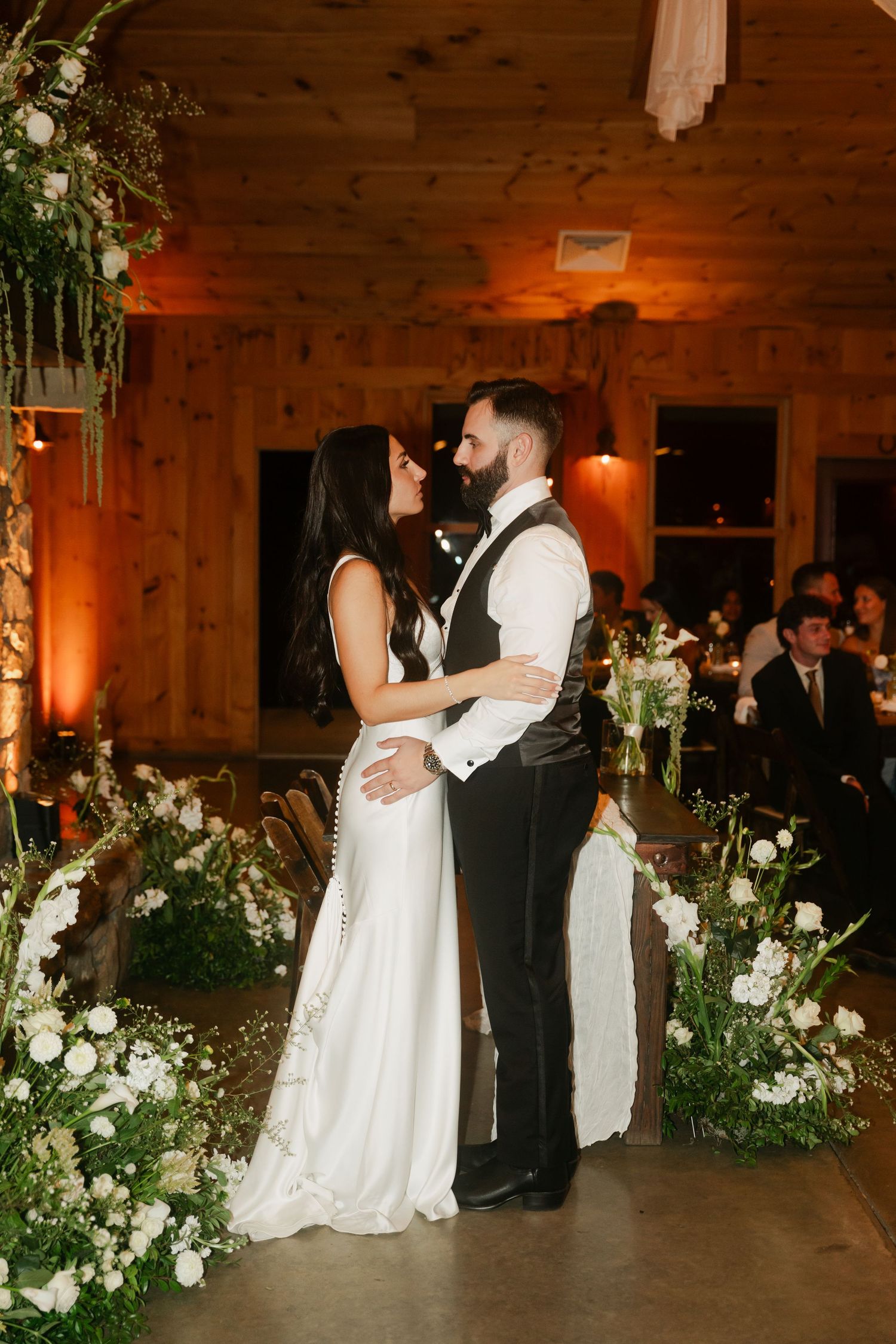 Bride and groom share romantic first dance moment in rustic barn wedding venue with white floral arch.
