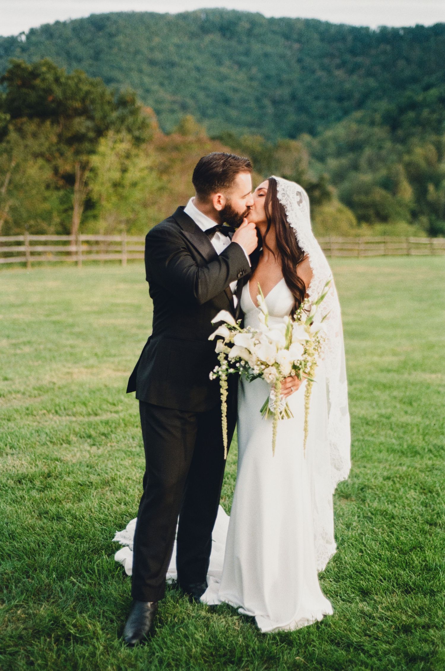 Bride and groom share a romantic kiss on a green lawn with mountain views on their wedding day.