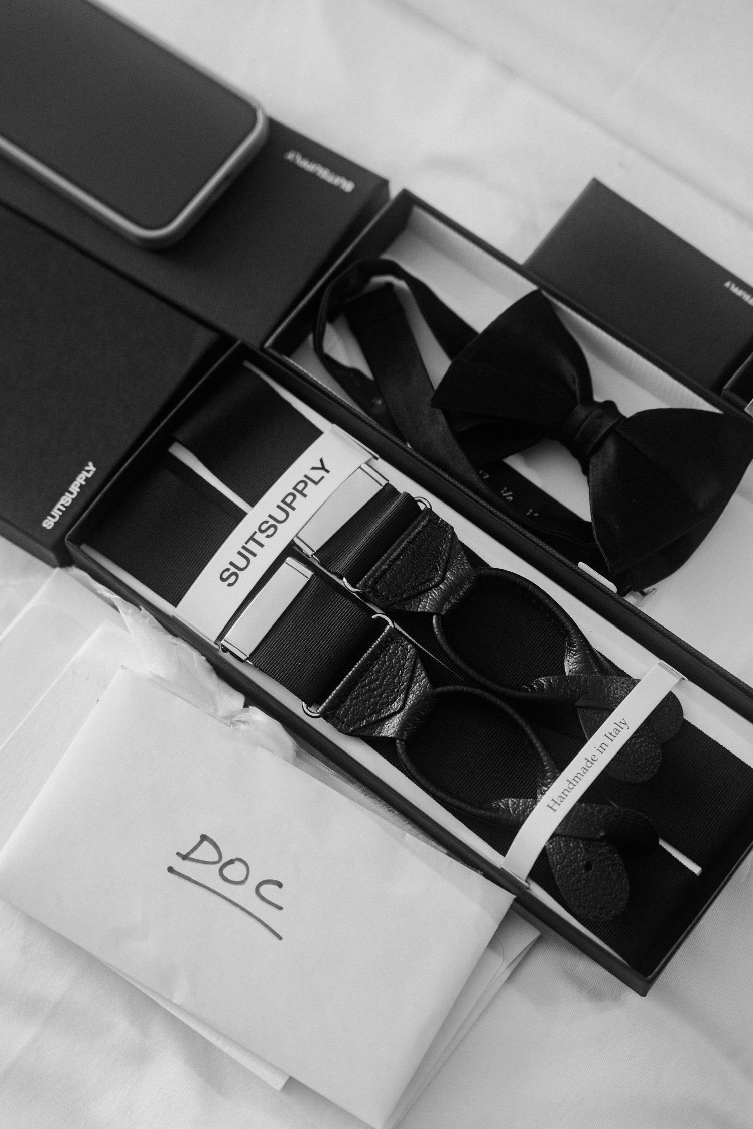 Black and white flat lay of SuitSupply suspenders, bow tie, and accessories in gift boxes for a wedding.