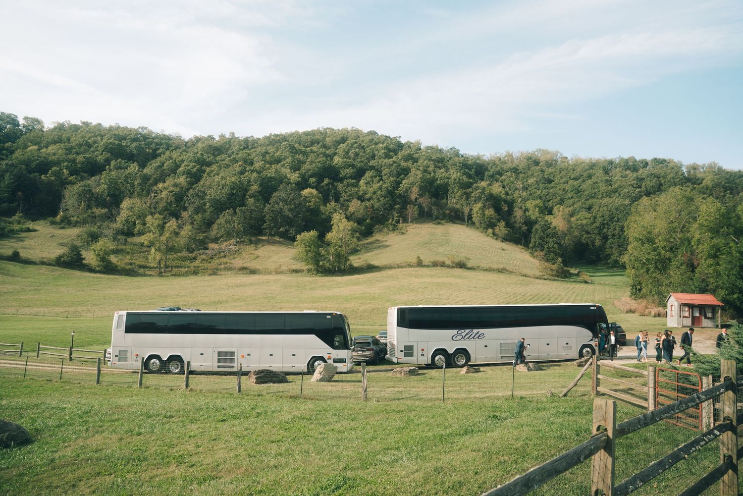 Two charter buses parked in a lush green valley field surrounded by forested hills at an outdoor wedding venue.