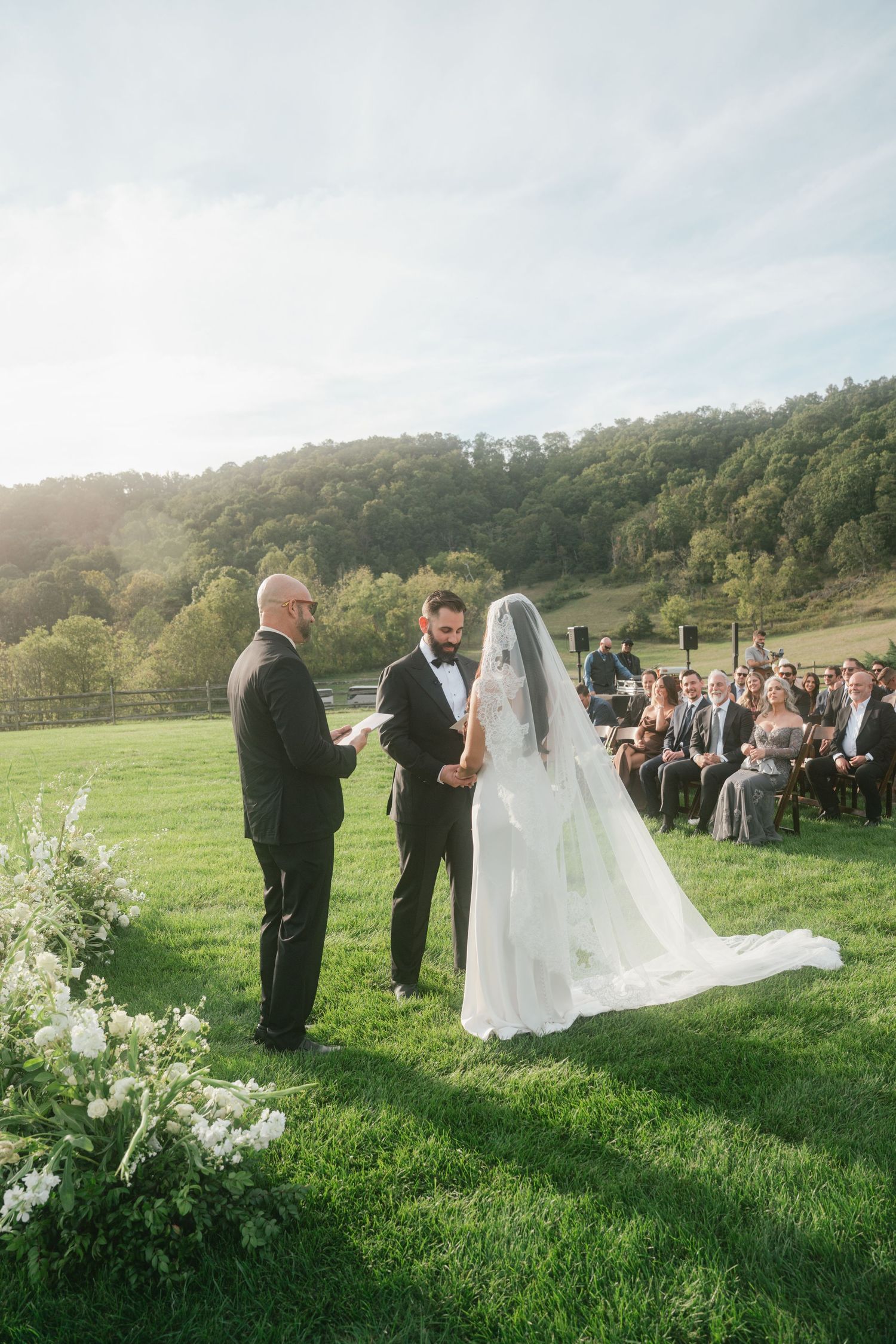 Bride and groom exchange vows outdoors on lush green lawn with rolling hills and seated wedding guests in background.