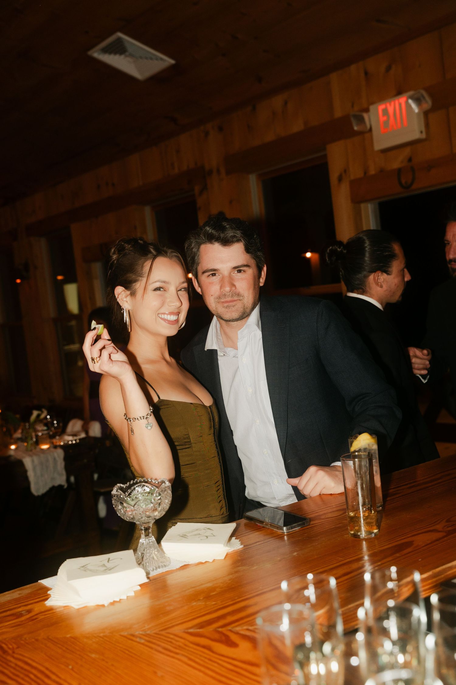Couple smiling together at wedding reception bar, woman holding cigar in rustic wood venue.