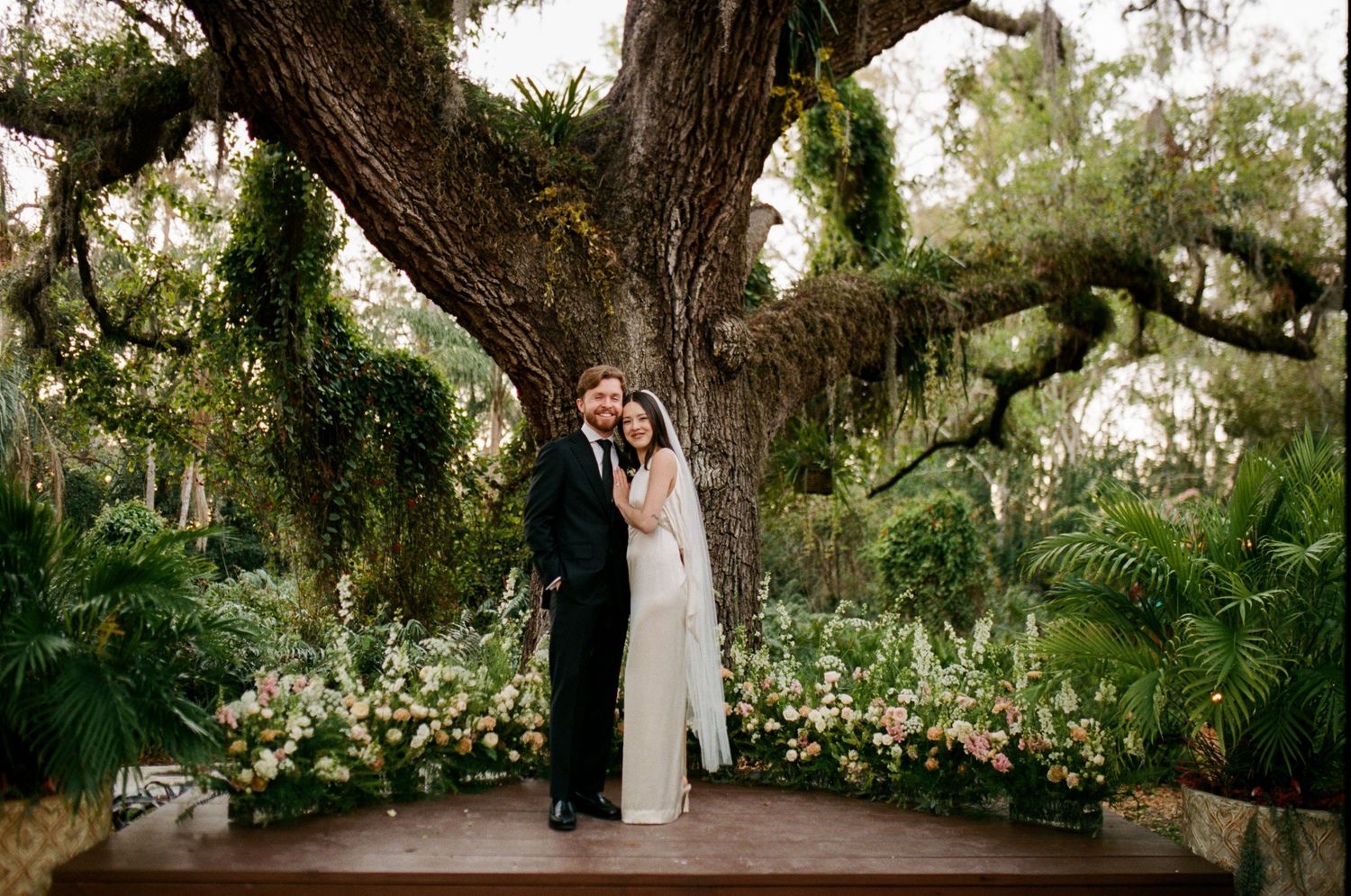 Bride and groom embrace under a grand oak tree surrounded by lush greenery and floral arrangements at an outdoor wedding.