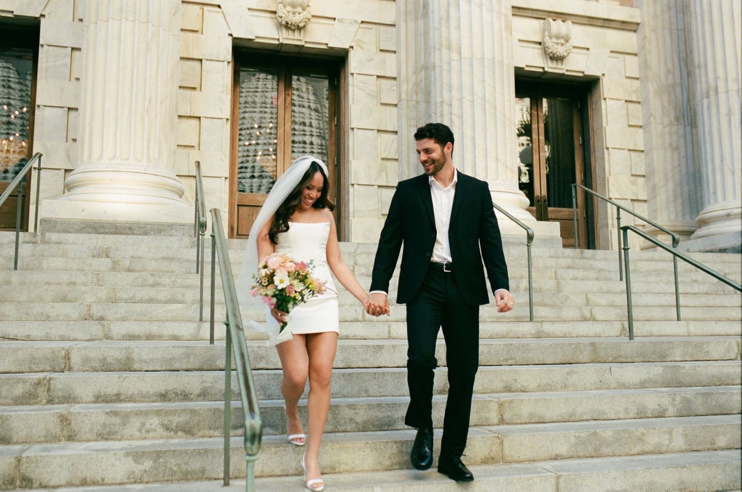 Newlywed couple holding hands walking down stone steps of a grand building, bride in short white dress with veil and bouquet.