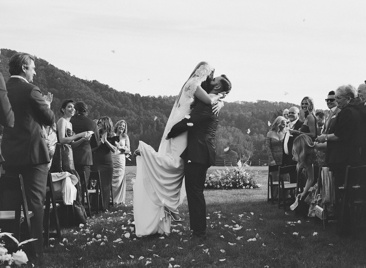 Black and white wedding photo of bride and groom sharing a first kiss while guests celebrate along the aisle.