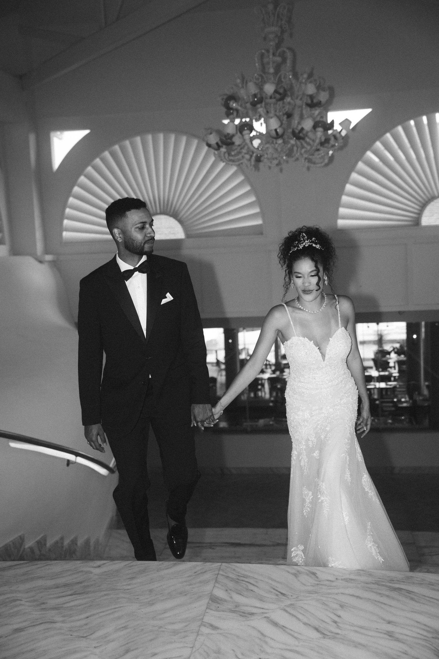 Elegant couple descending stairs in formal attire under decorative fan windows.