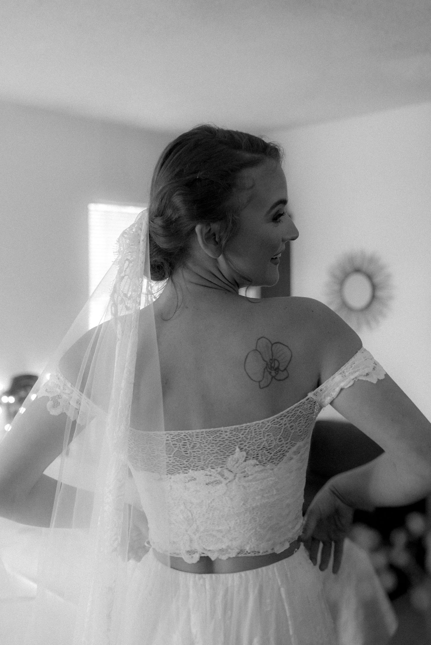 Back view of a bride in a white lace dress showing an orchid tattoo and flowing veil.
