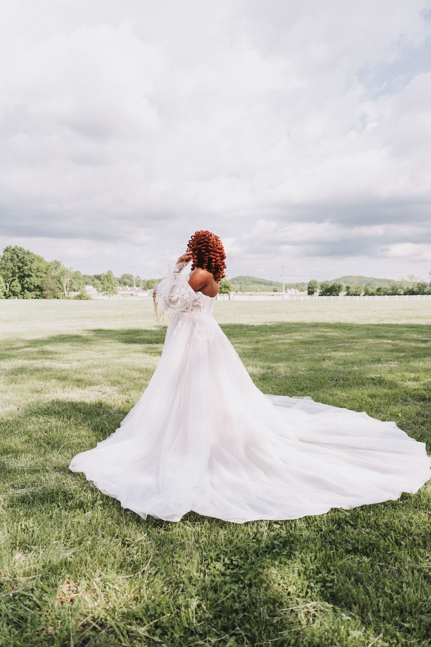Bride in flowing white gown spins on grassy field under cloudy sky.