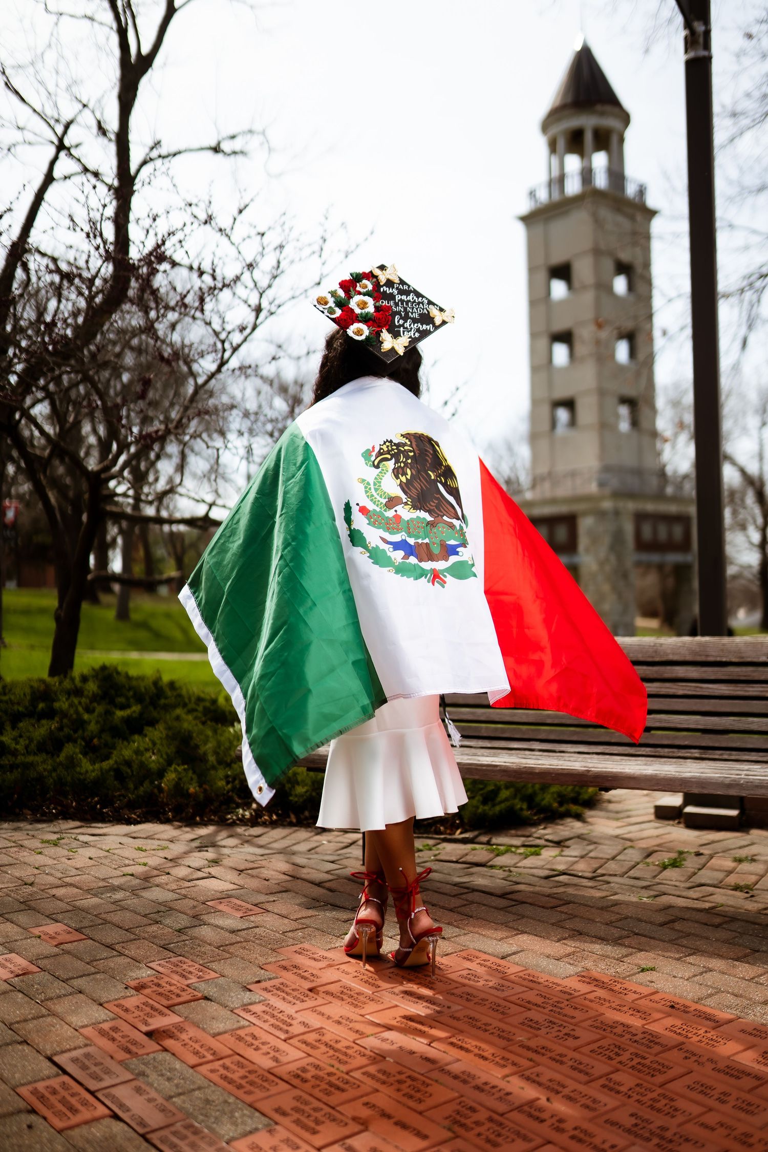 Graduate wearing Mexican flag cape stands in front of bell tower on brick pathway.
