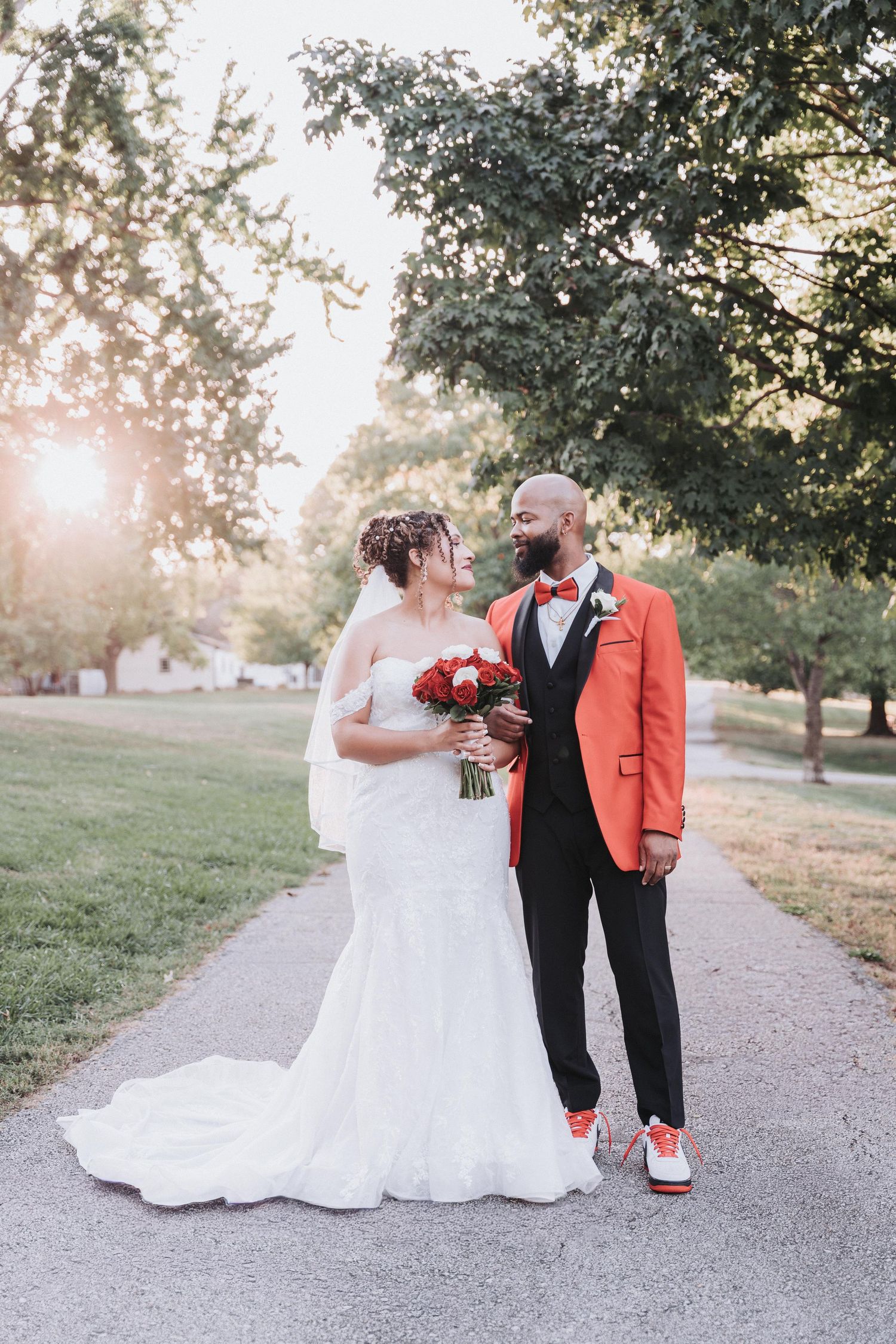 A couple in wedding attire poses on a path at sunset, with the bride in white and groom wearing a coral suit jacket.