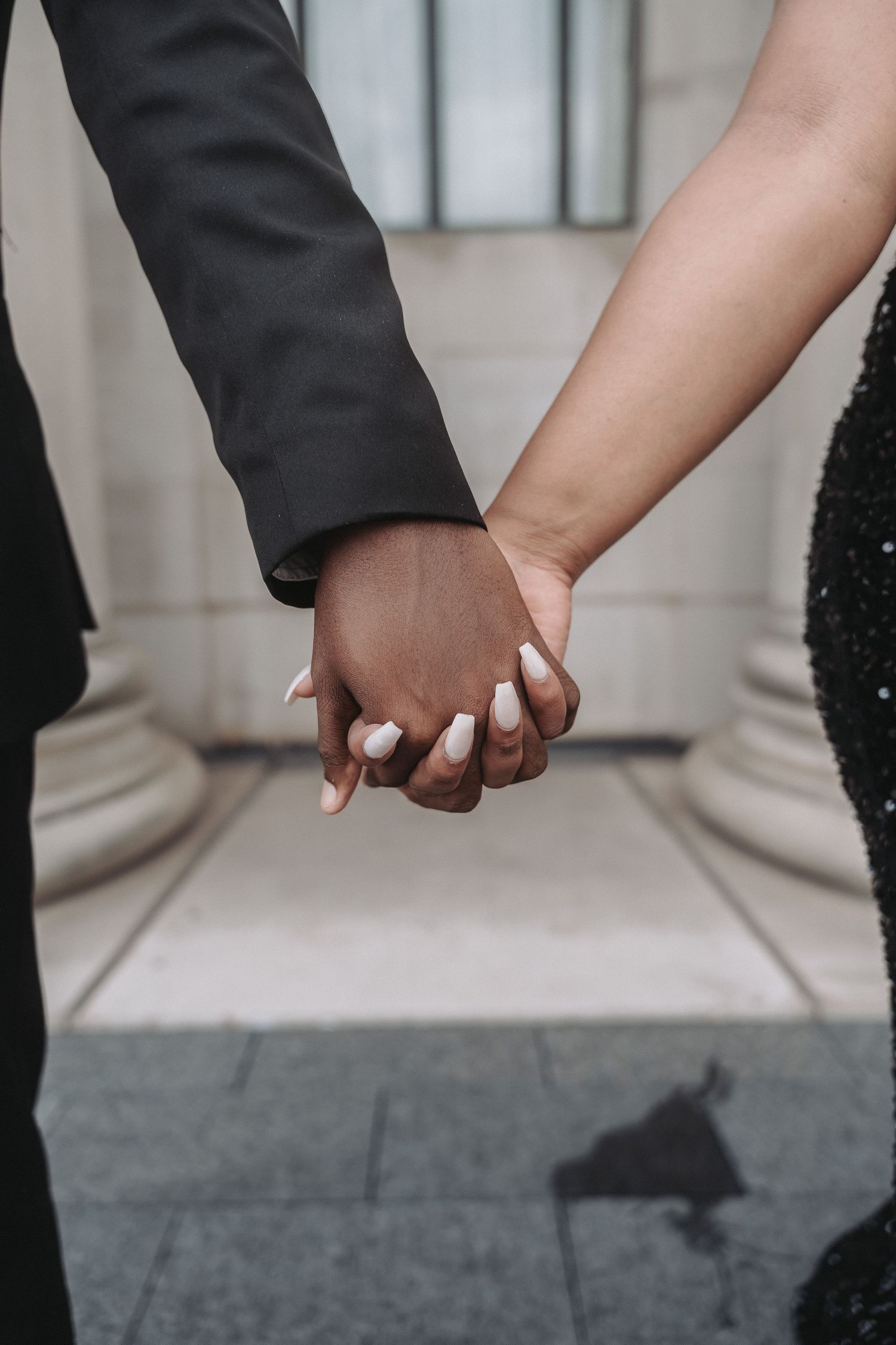 Close-up of interracial couple holding hands romantically against a light stone building backdrop.