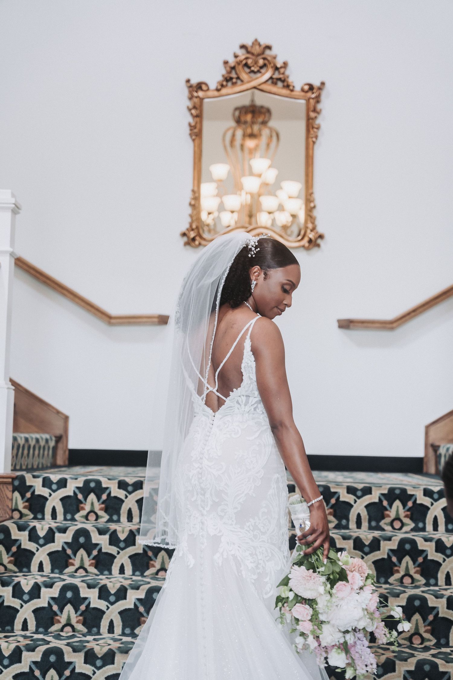 Bride in fitted lace gown poses on ornate staircase with vintage mirror and floral bouquet.
