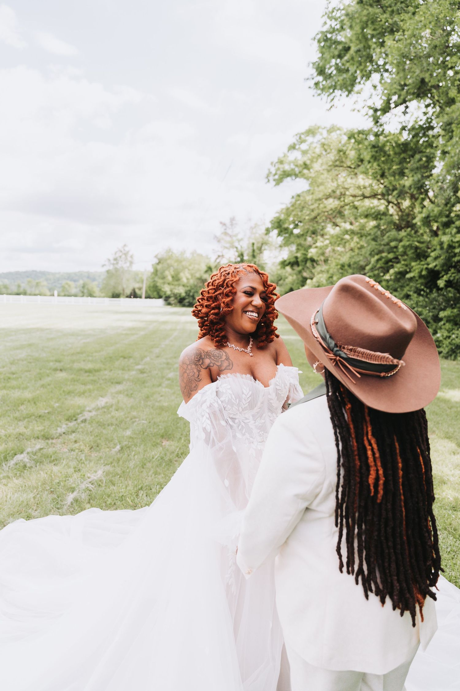 Two people in white outfits share a joyful moment together in a scenic outdoor setting with green trees and grass.