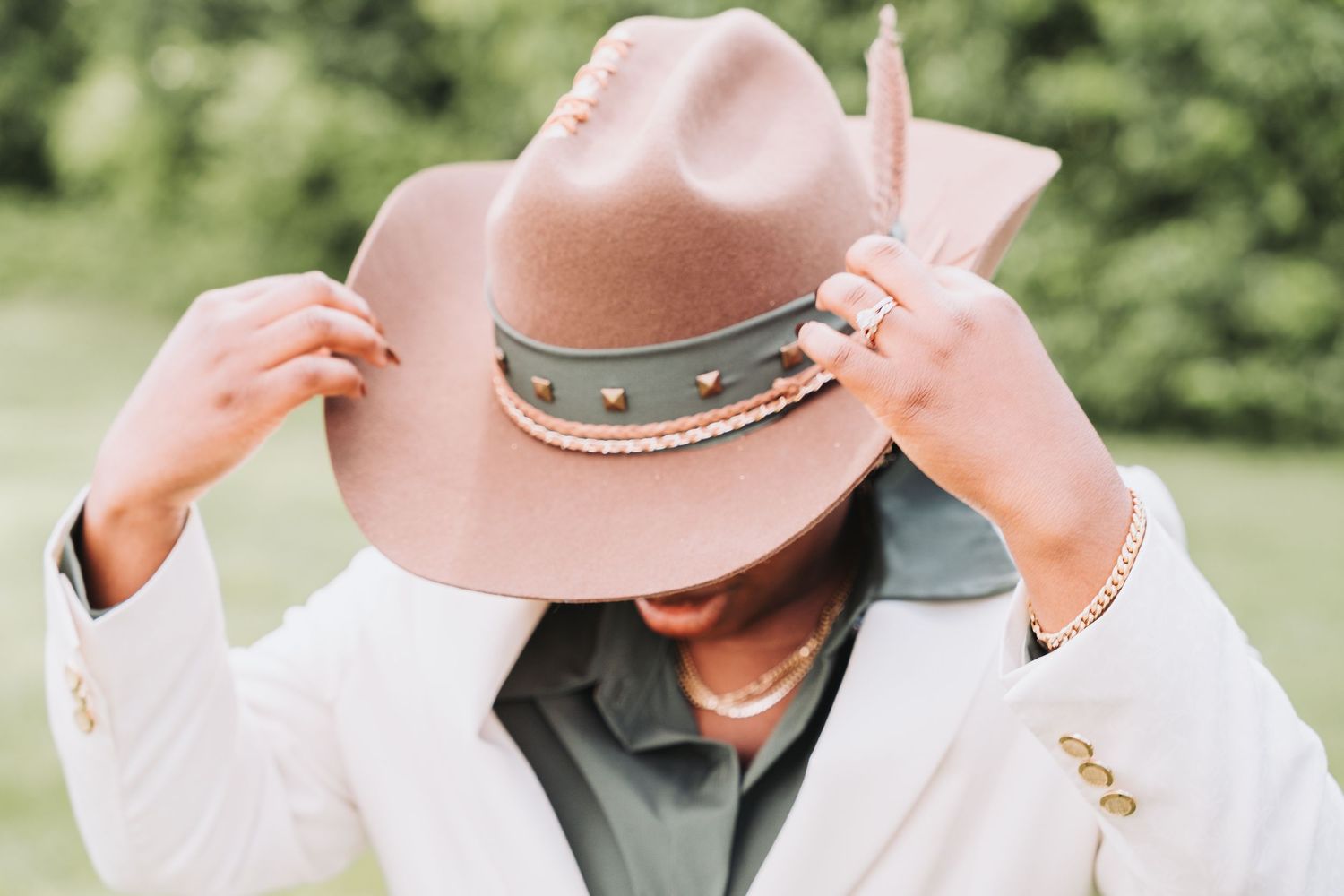 Someone wearing a white blazer adjusts a pink felt cowboy hat with studded band detail against blurred green background.