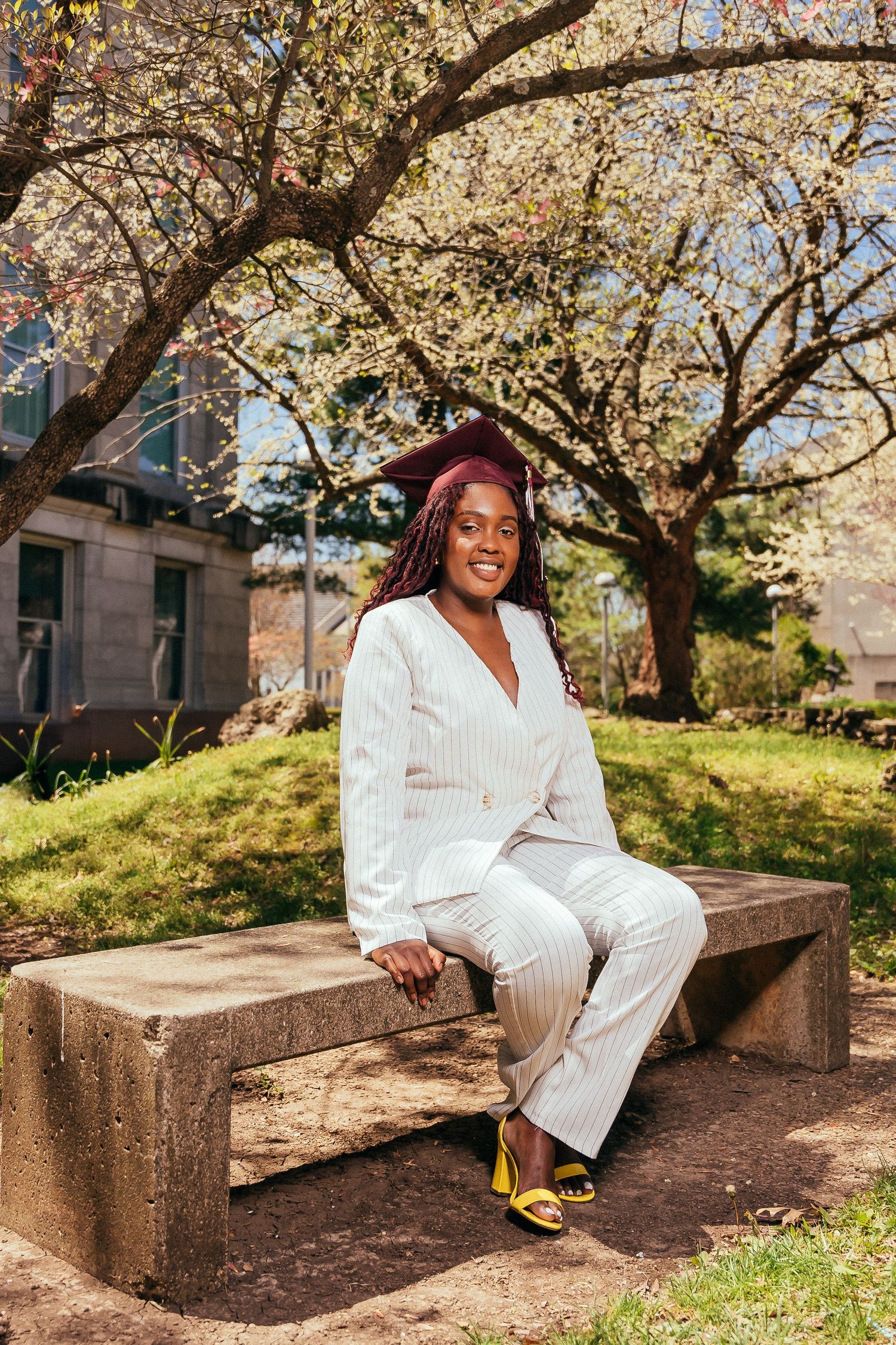 Graduate in white outfit sits on concrete bench under blooming cherry trees.