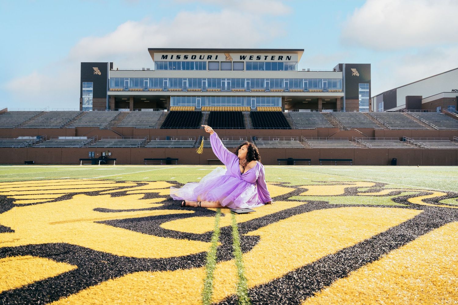 Graduate in flowing purple dress poses on football field with stadium in background.