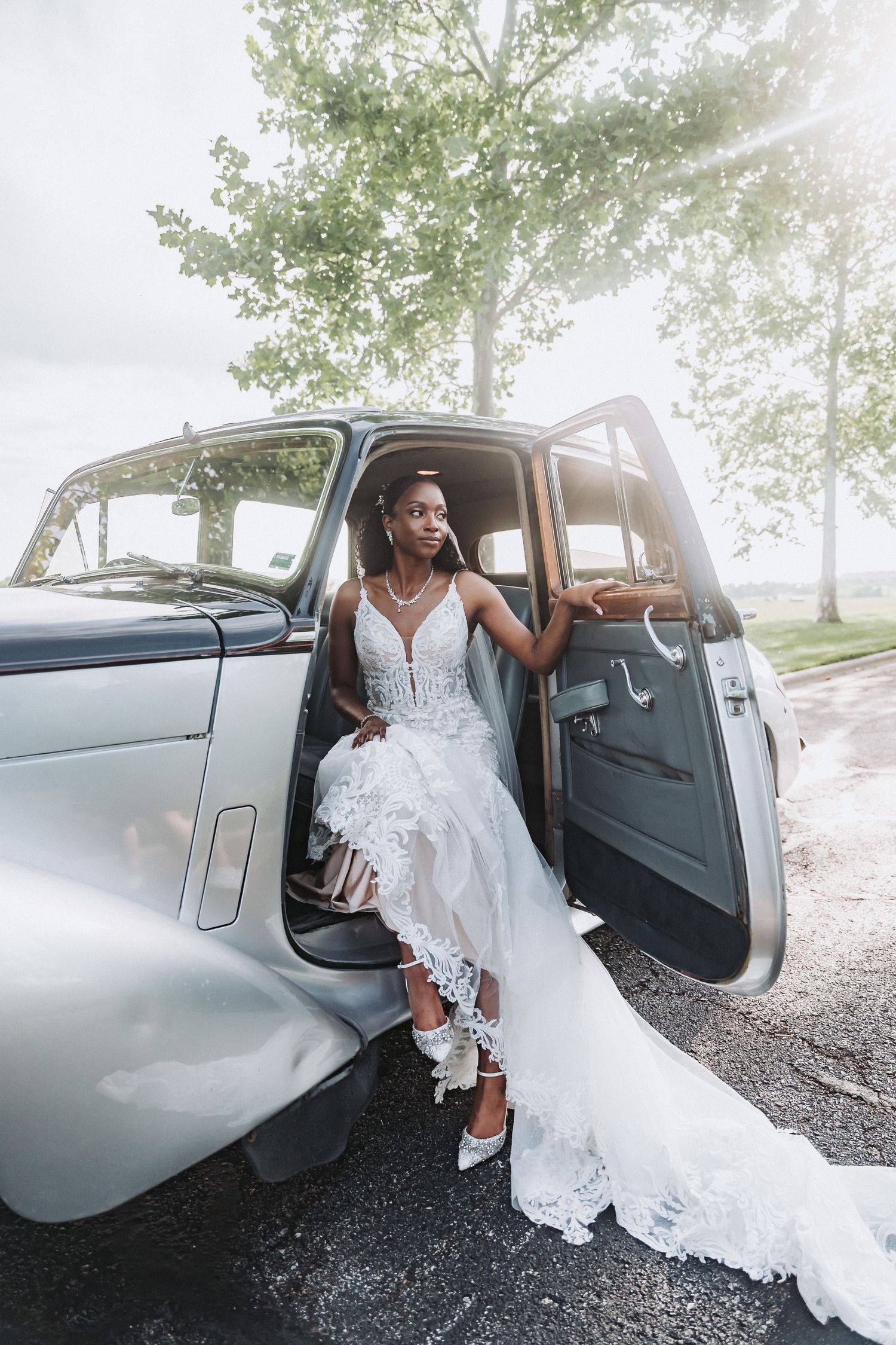 A bride in a lace gown poses elegantly from inside a vintage car with her dress flowing out.