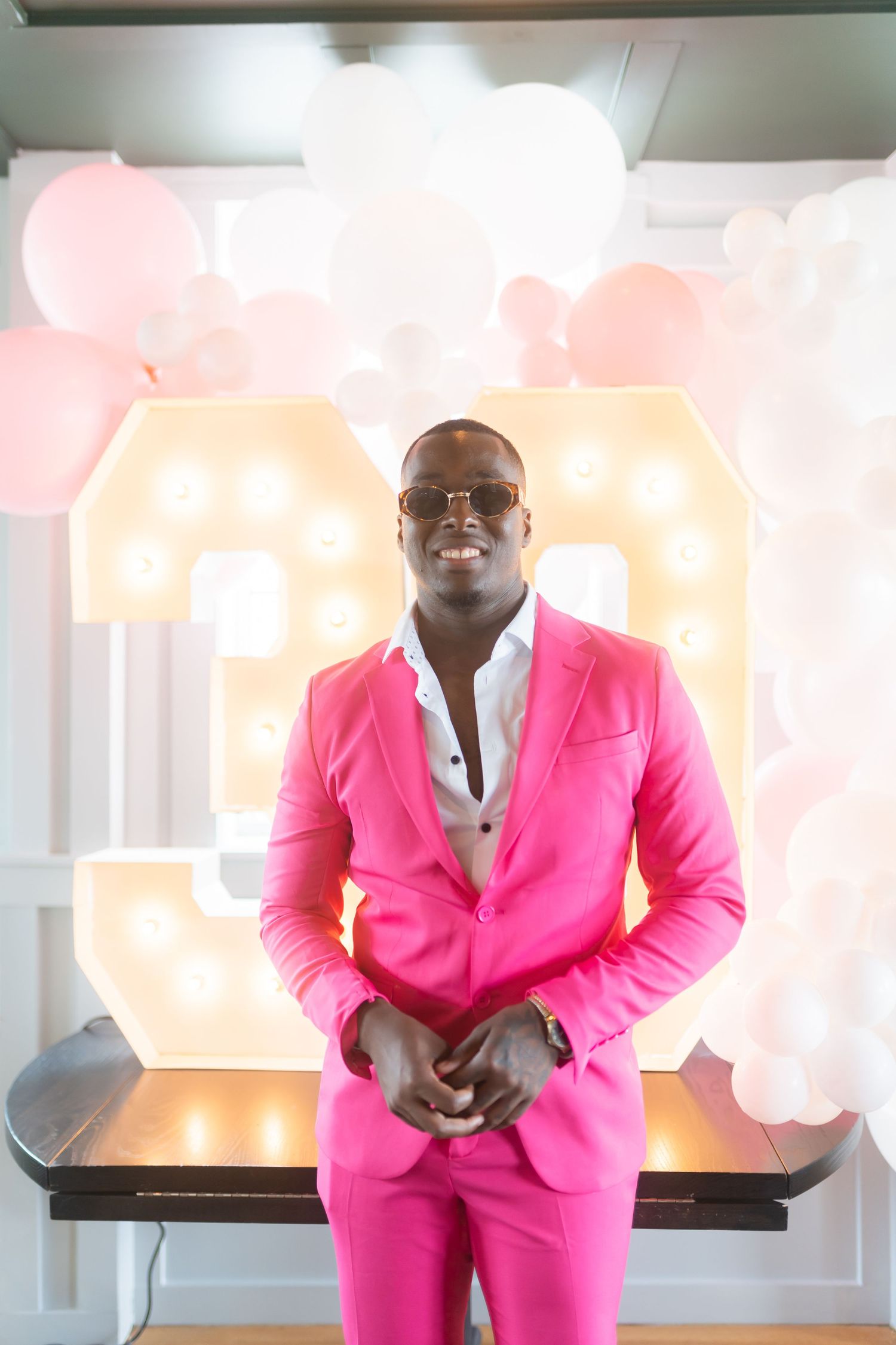 Person wearing bright pink suit stands in front of illuminated number display with balloons.