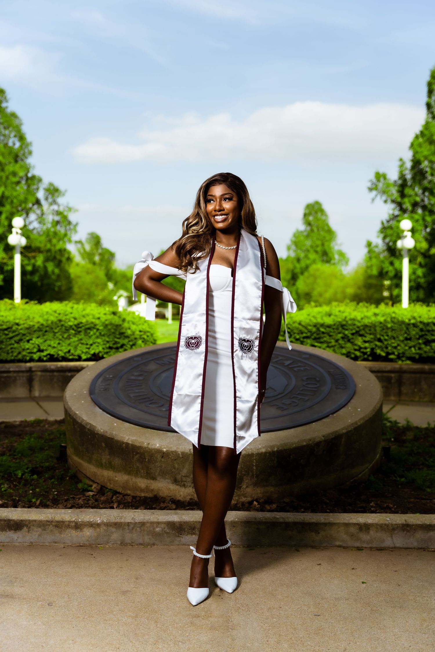 Graduate in white dress and academic stole poses by circular stone fountain.