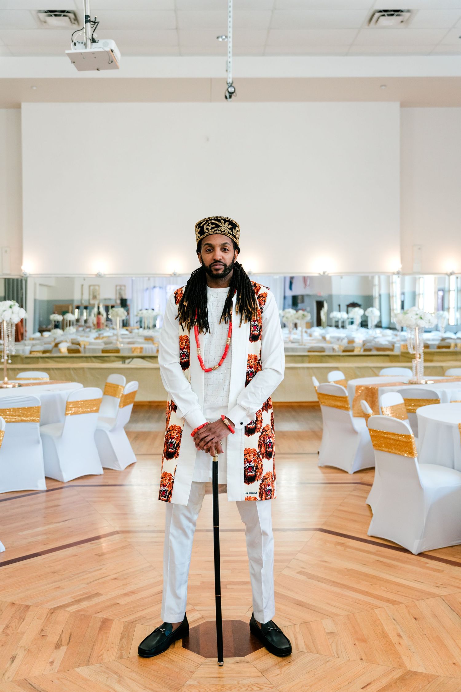 Groom in white suit with African-inspired accessories and crown stands in reception venue.