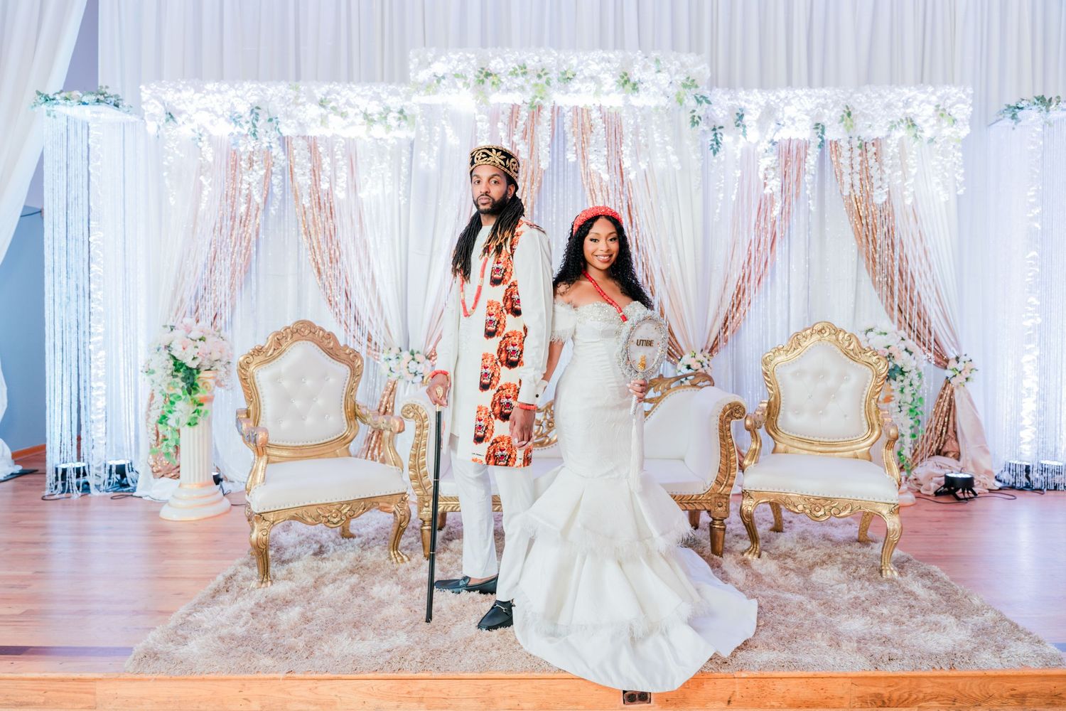 Wedding couple poses in traditional attire against elegant white and gold backdrop with thrones.