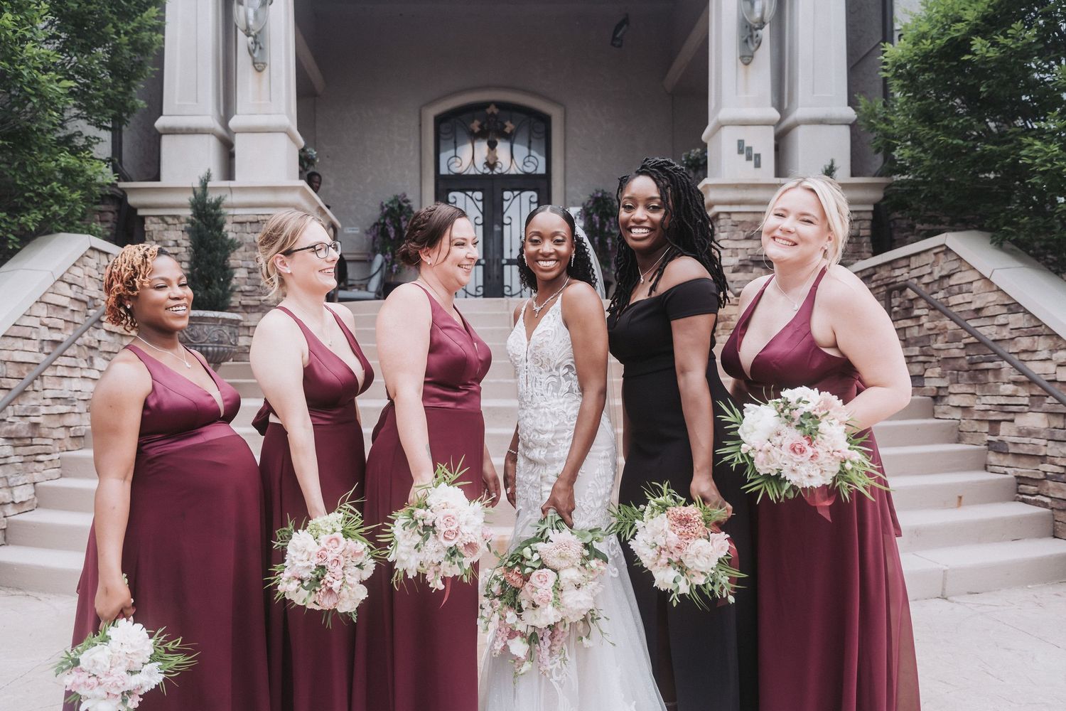 Bridal party in burgundy dresses poses on stone steps with white floral bouquets.