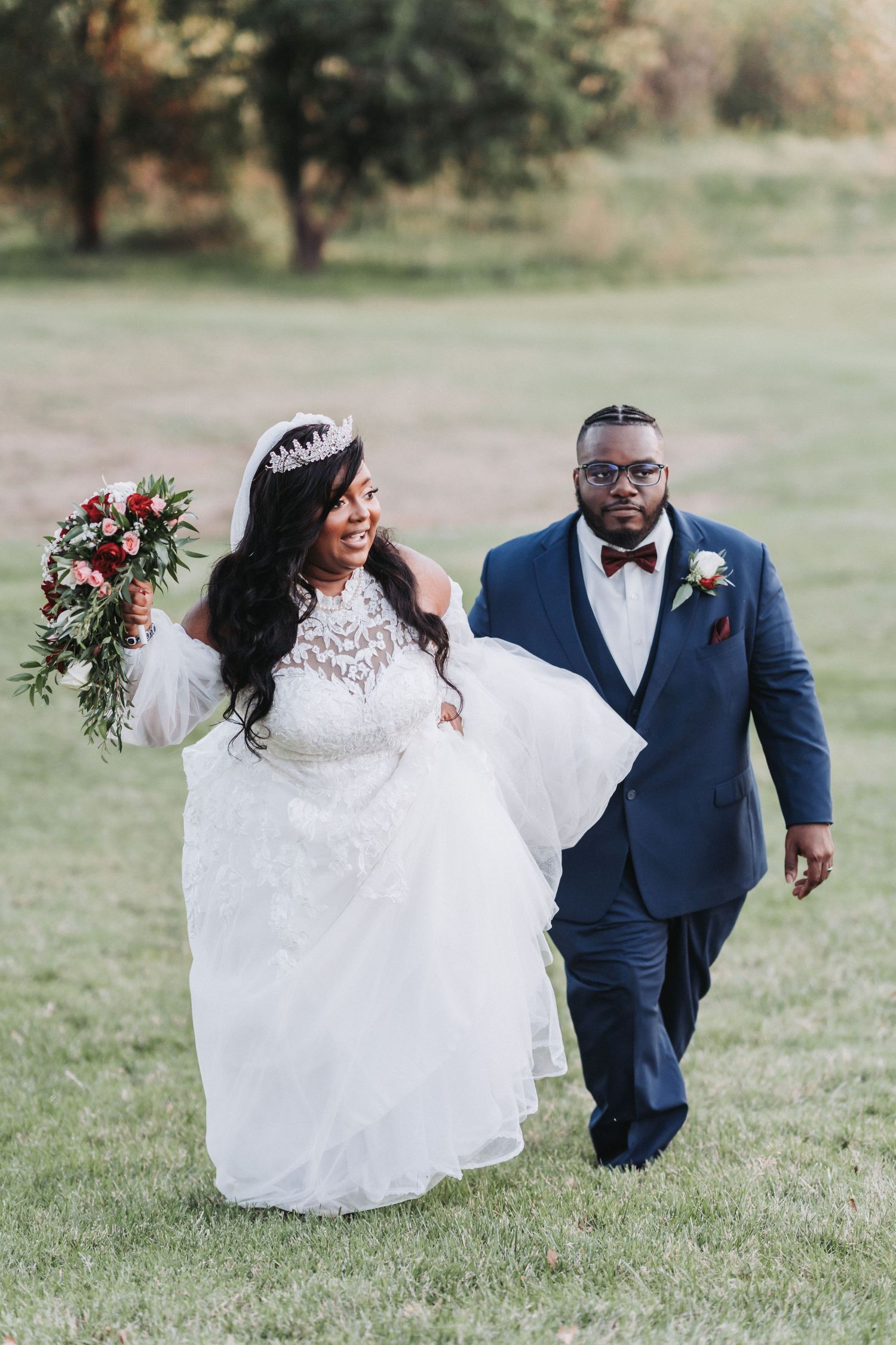 Bride in lace dress and groom in navy suit walk together across grass holding bouquet.