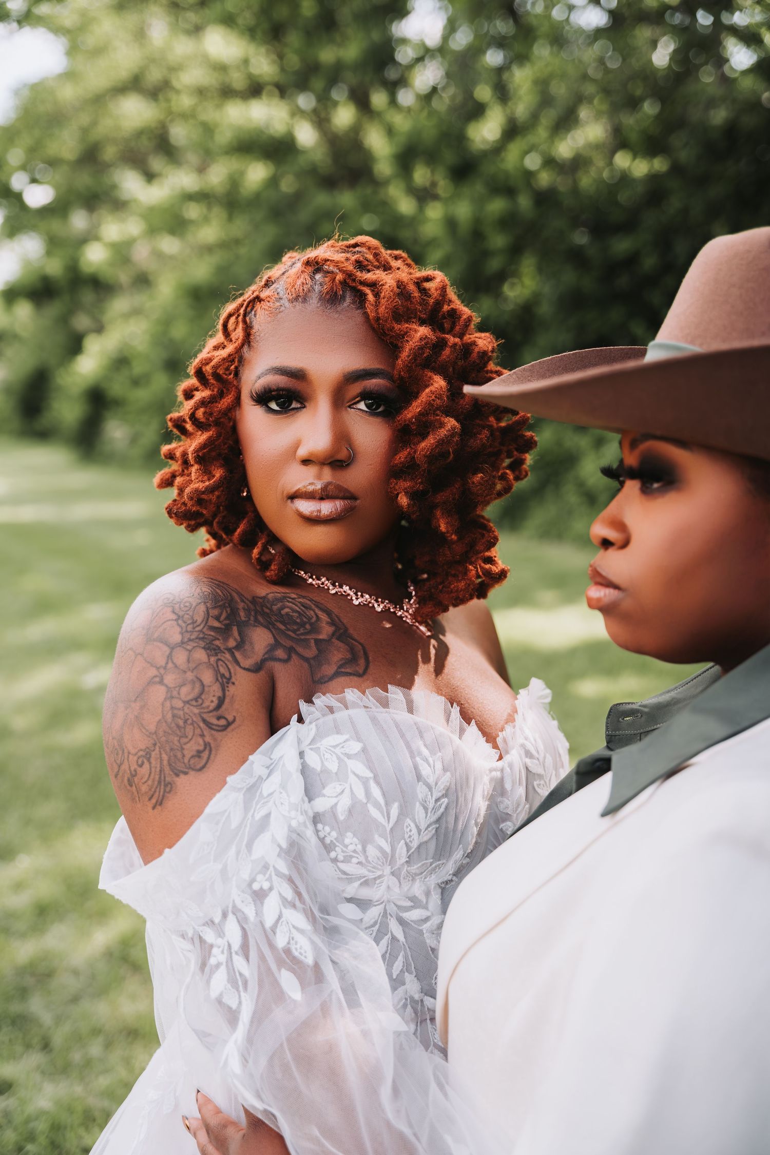 Two people in wedding attire pose outdoors, one wearing a white lace dress with tattoos and another in a brown hat.