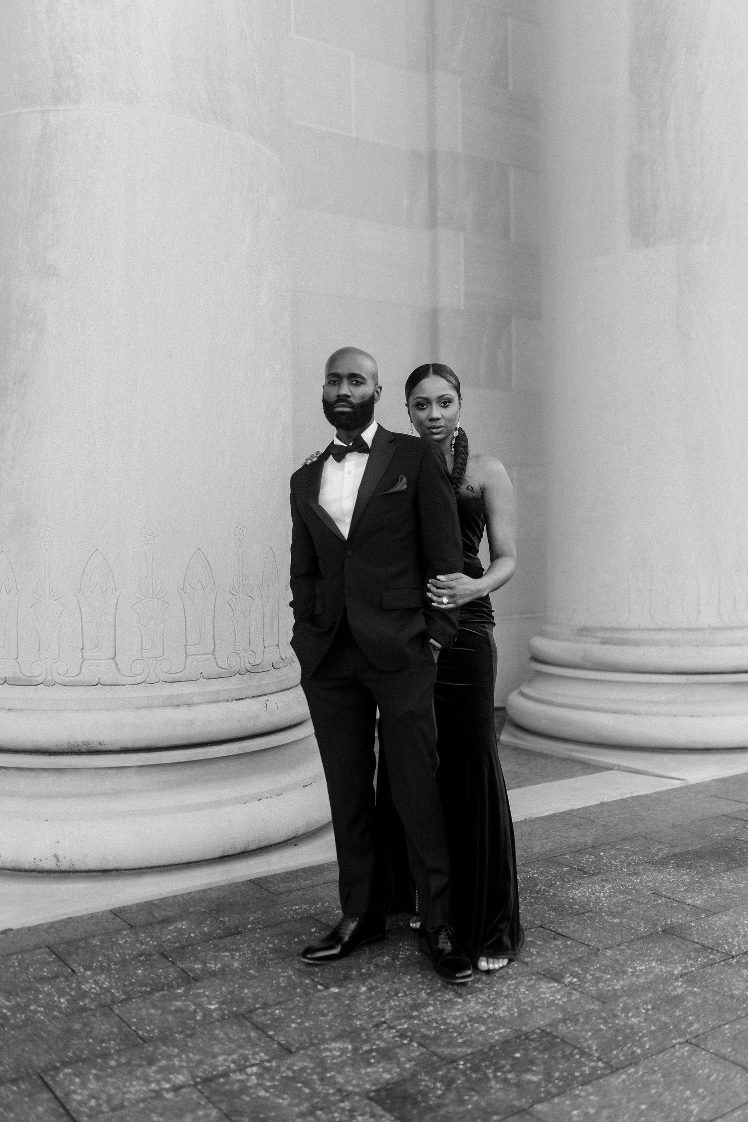Black and white portrait of formally dressed couple standing between classic architectural columns.