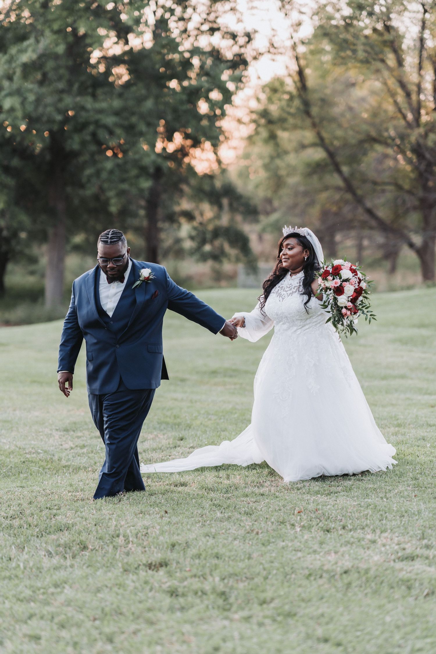 Romantic wedding couple walks hand in hand across a sunlit lawn with a floral bouquet.