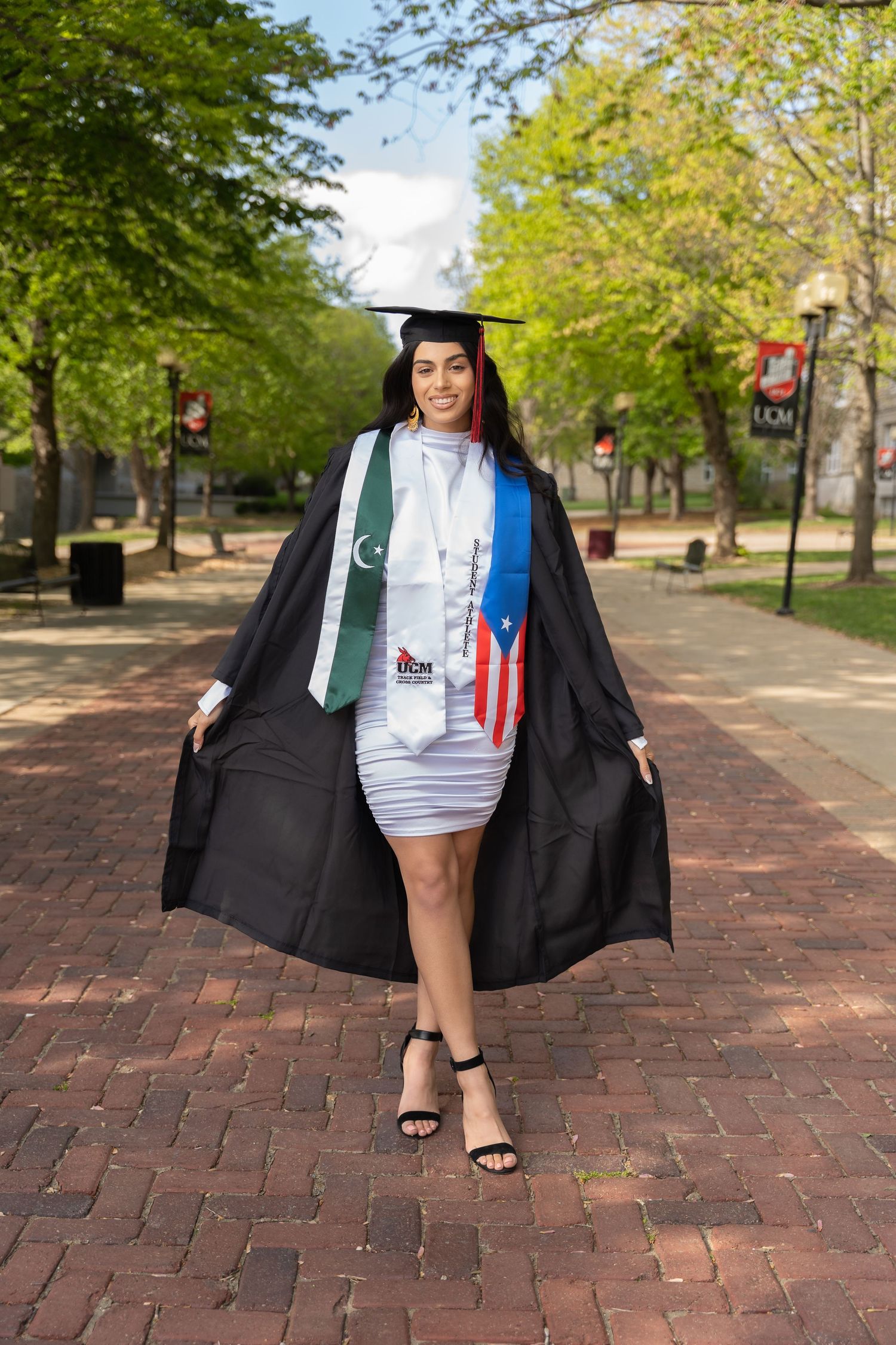 Graduate in black cap and gown with colorful stoles stands on brick pathway surrounded by green trees.