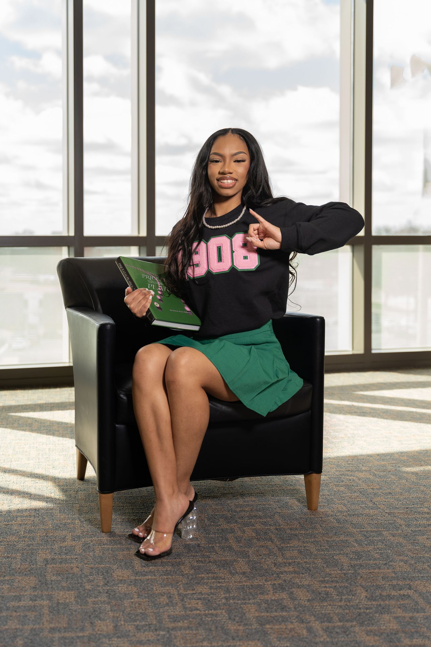 A professional woman in a 306 sweatshirt poses cheerfully on a black armchair near floor-to-ceiling windows.