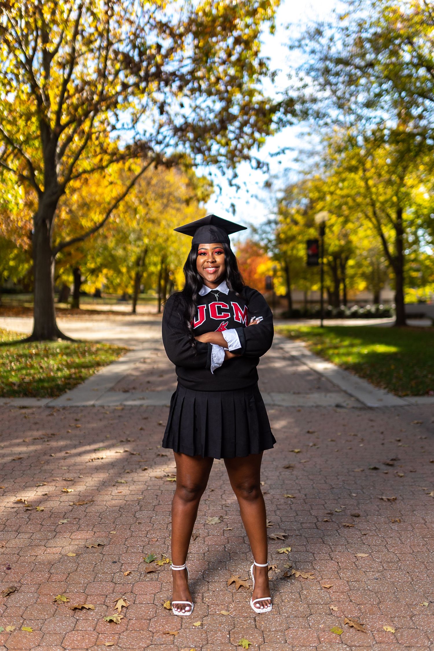 Graduate in black cap and gown poses confidently on brick pathway surrounded by fall foliage.