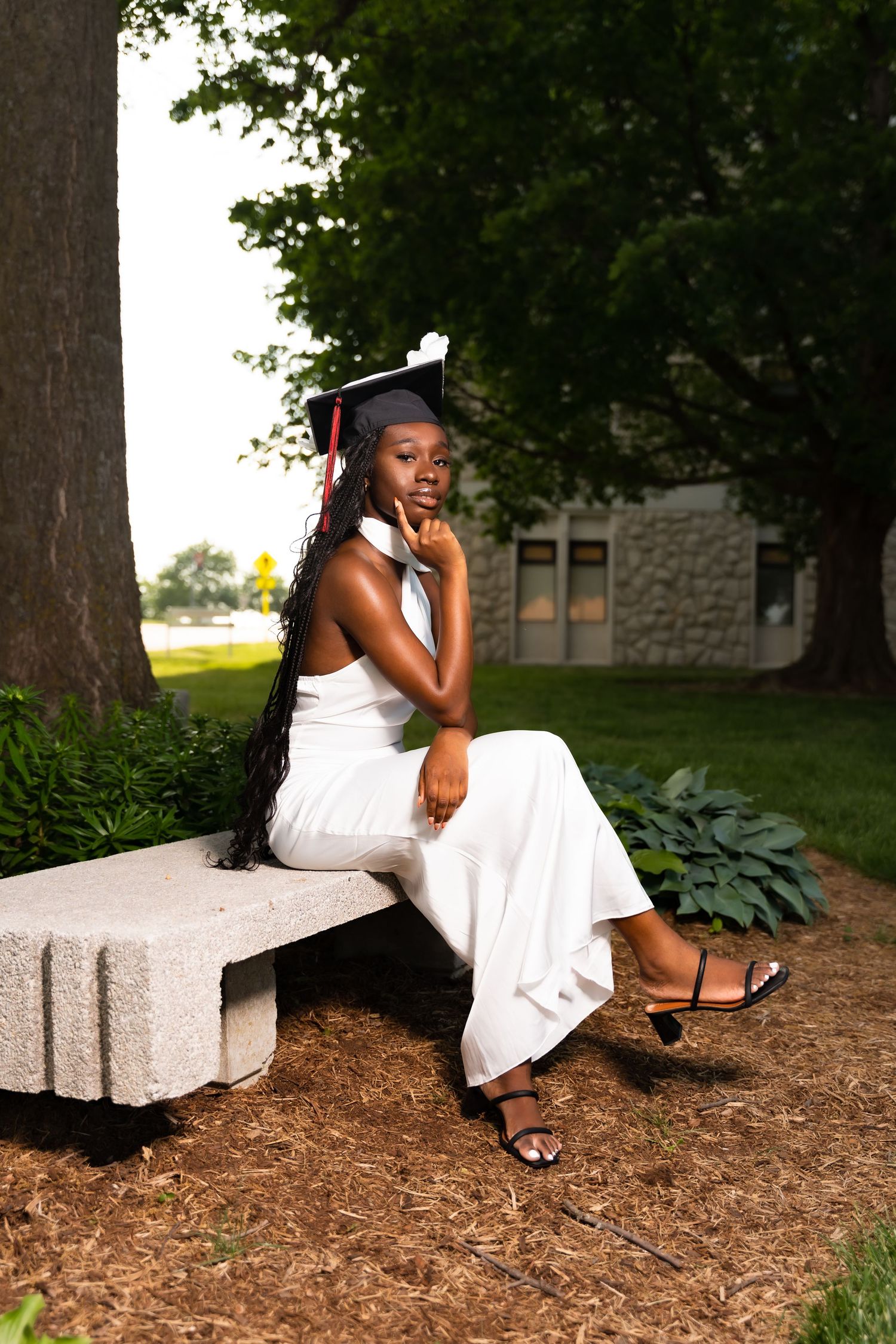 Graduate in white dress and cap poses elegantly on stone bench under tree.