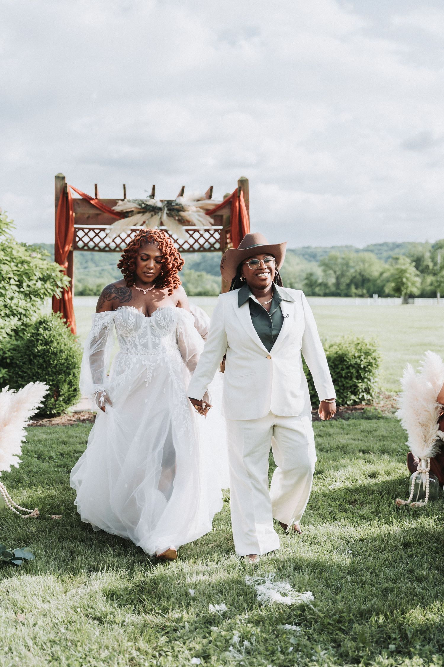 A couple walks hand in hand on their wedding day, wearing white attire in front of a rustic wooden arch outdoors.