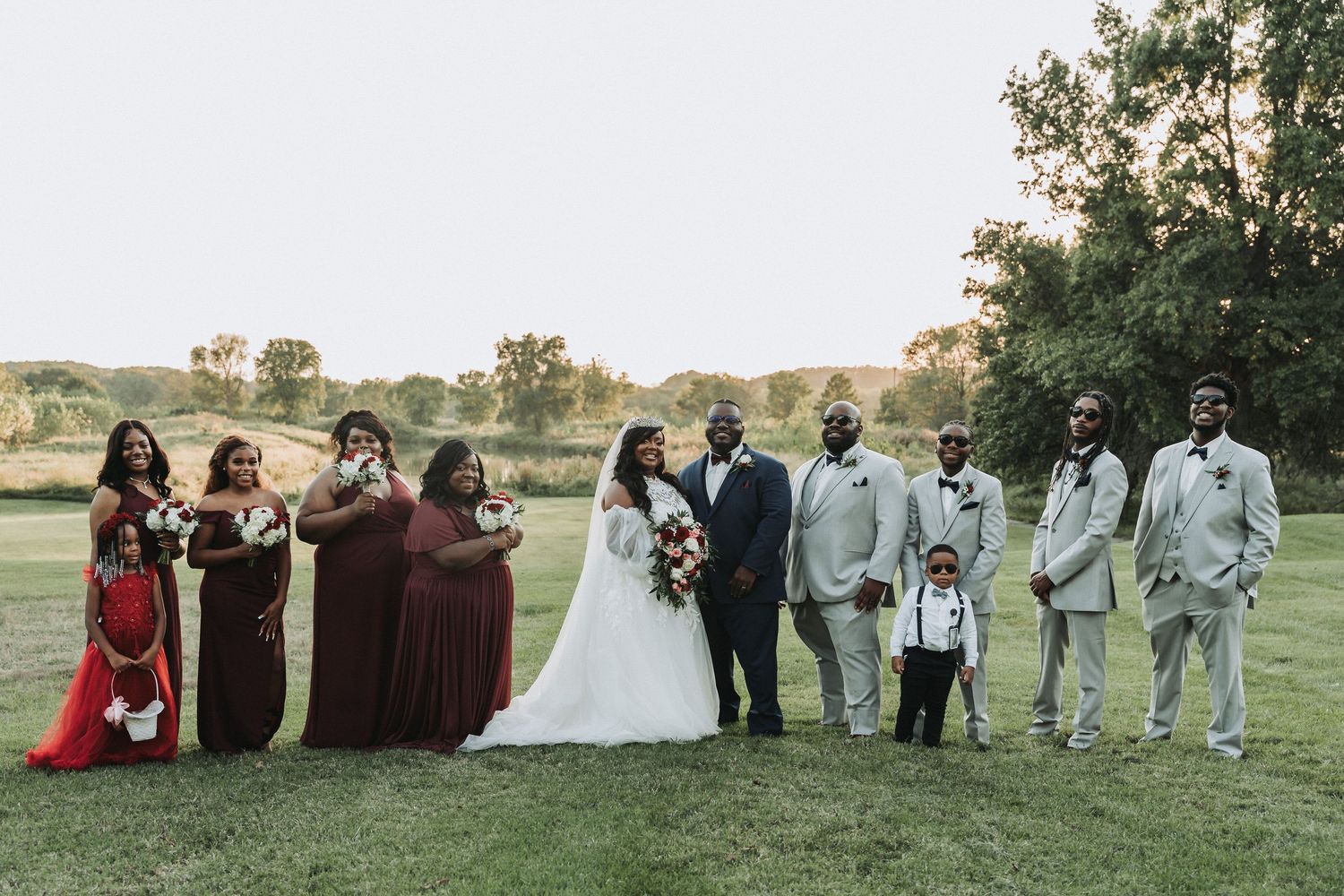 Wedding party in burgundy and gray attire poses together on a grassy lawn at sunset.
