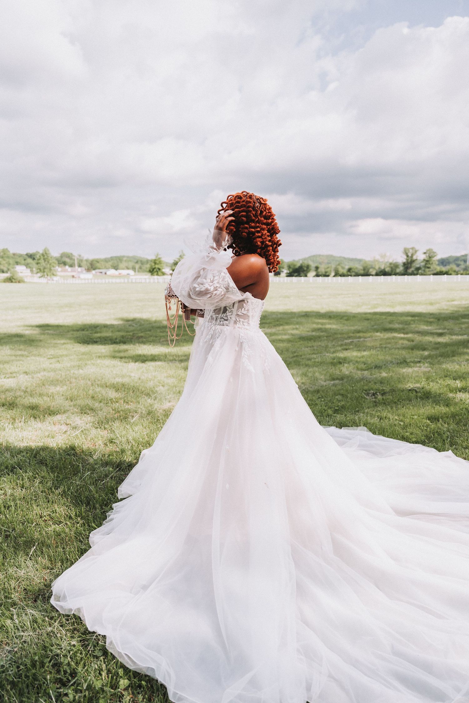 A bride in a flowing white gown twirls in a sunny field with dramatic cloudy sky.