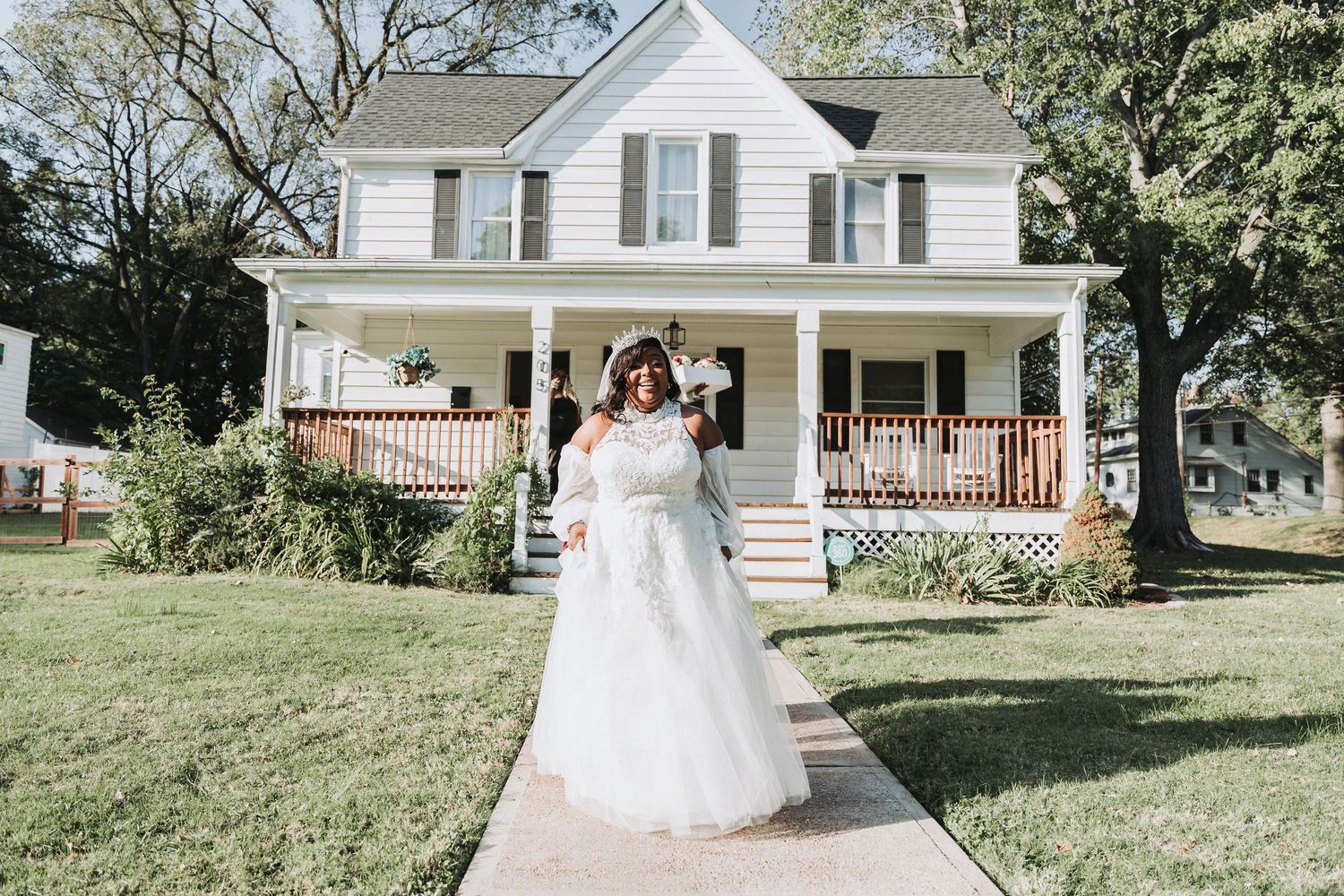 A bride in a white wedding dress poses in front of a classic white farmhouse with wraparound porch.