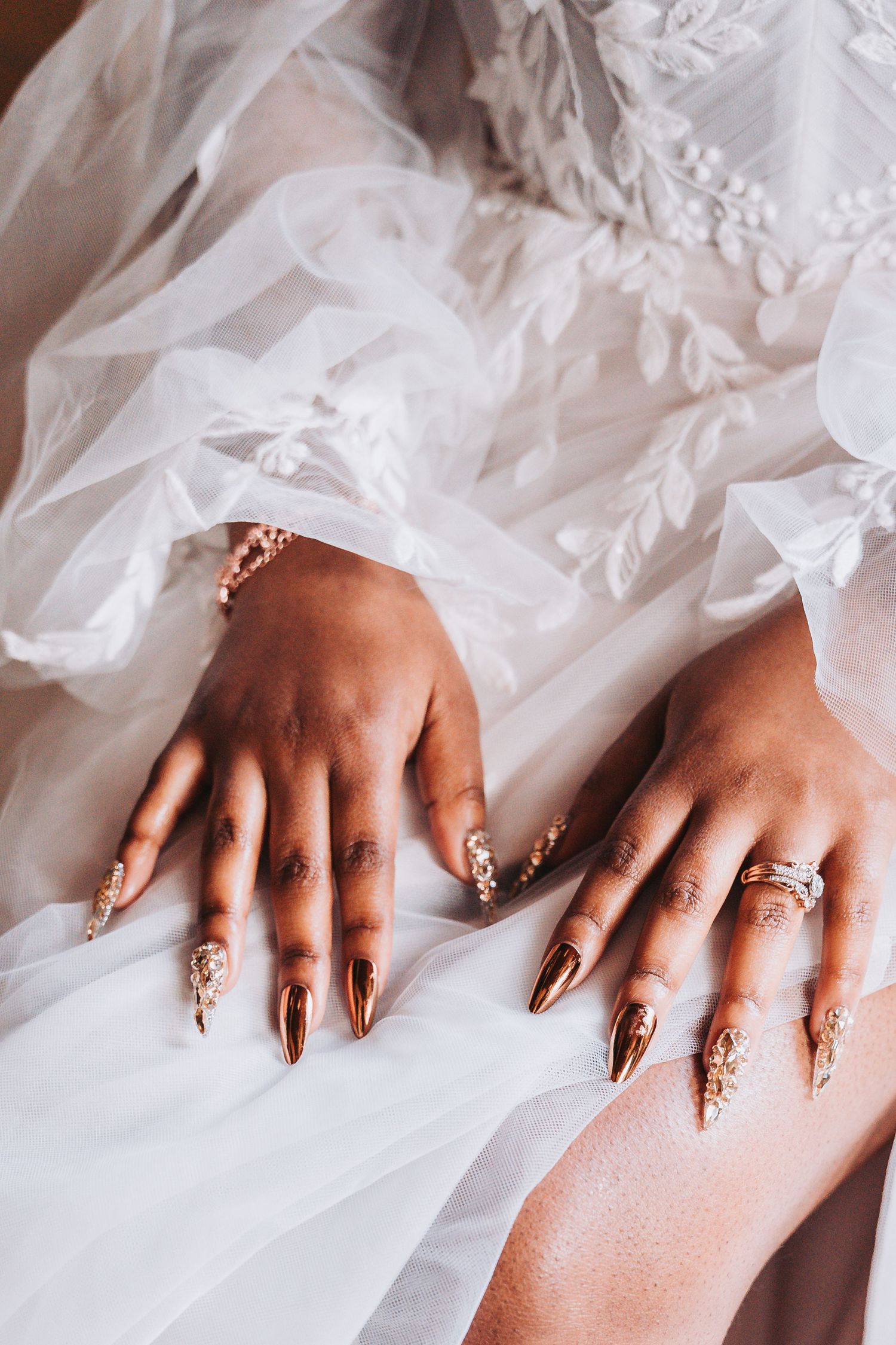 Ornate gold and crystal nail art adorns hands resting on white tulle wedding dress fabric.