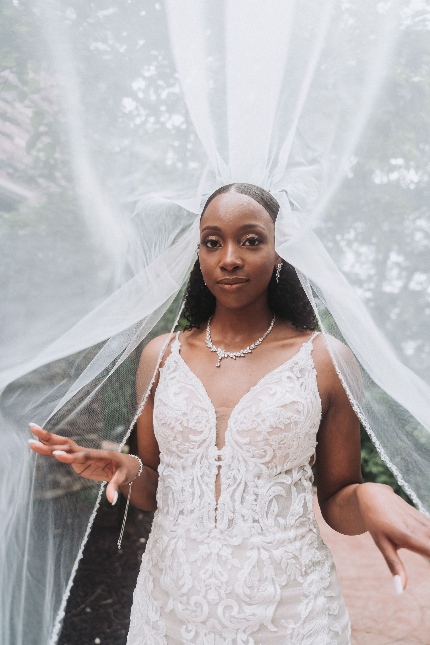 Bride in plunging neckline lace gown poses playfully with flowing veil outdoors.