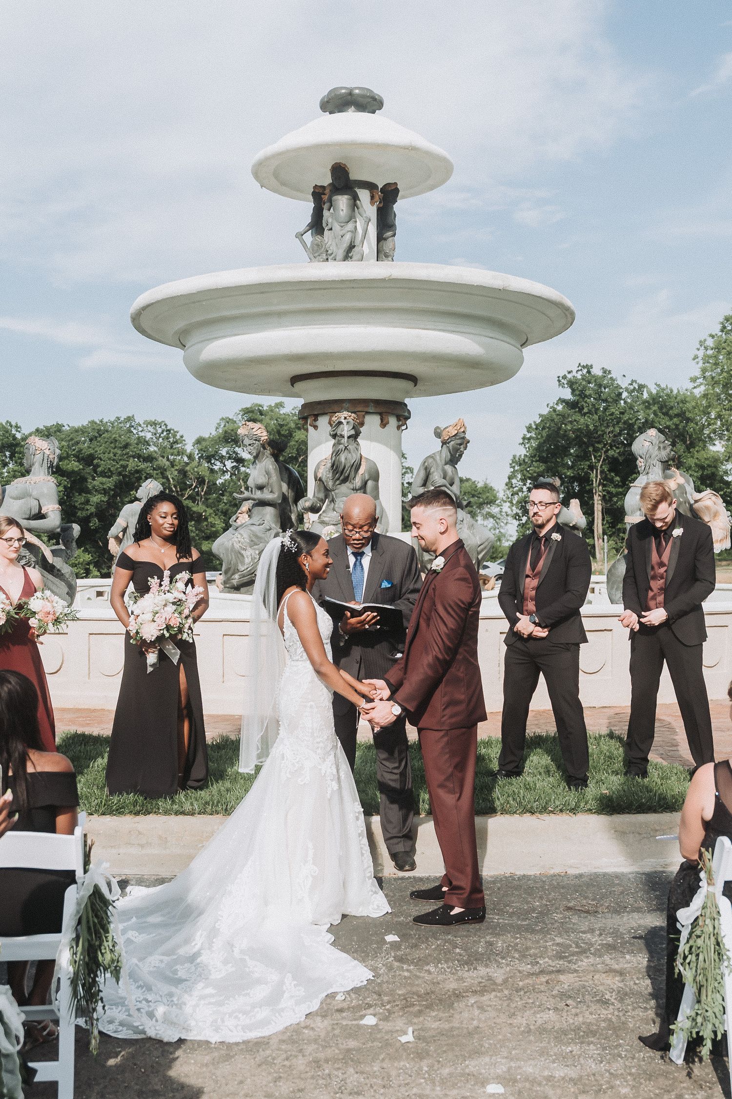 An outdoor wedding ceremony takes place in front of an ornate white fountain with guests looking on.