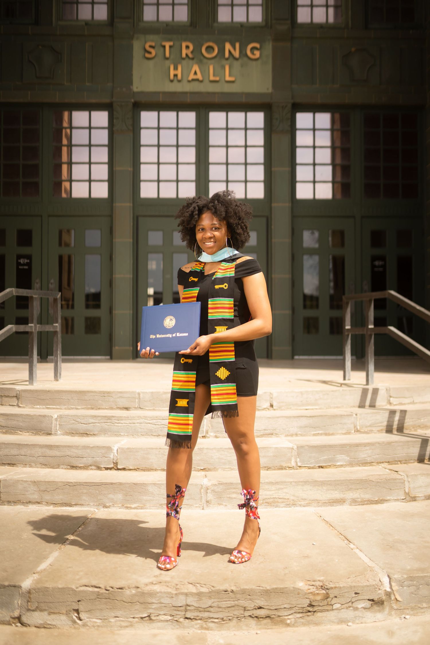 Graduate in traditional kente stole poses on steps of Strong Hall with diploma.