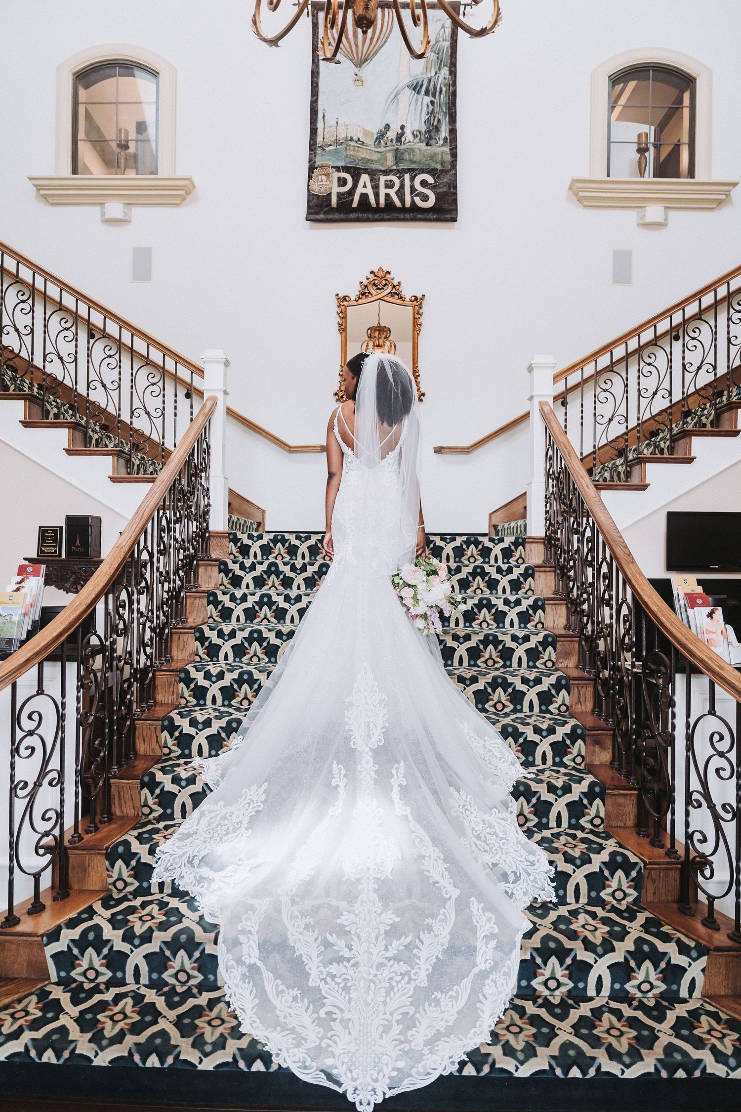 Bride in lace gown poses on grand staircase with Paris-themed decor and ornate railings.