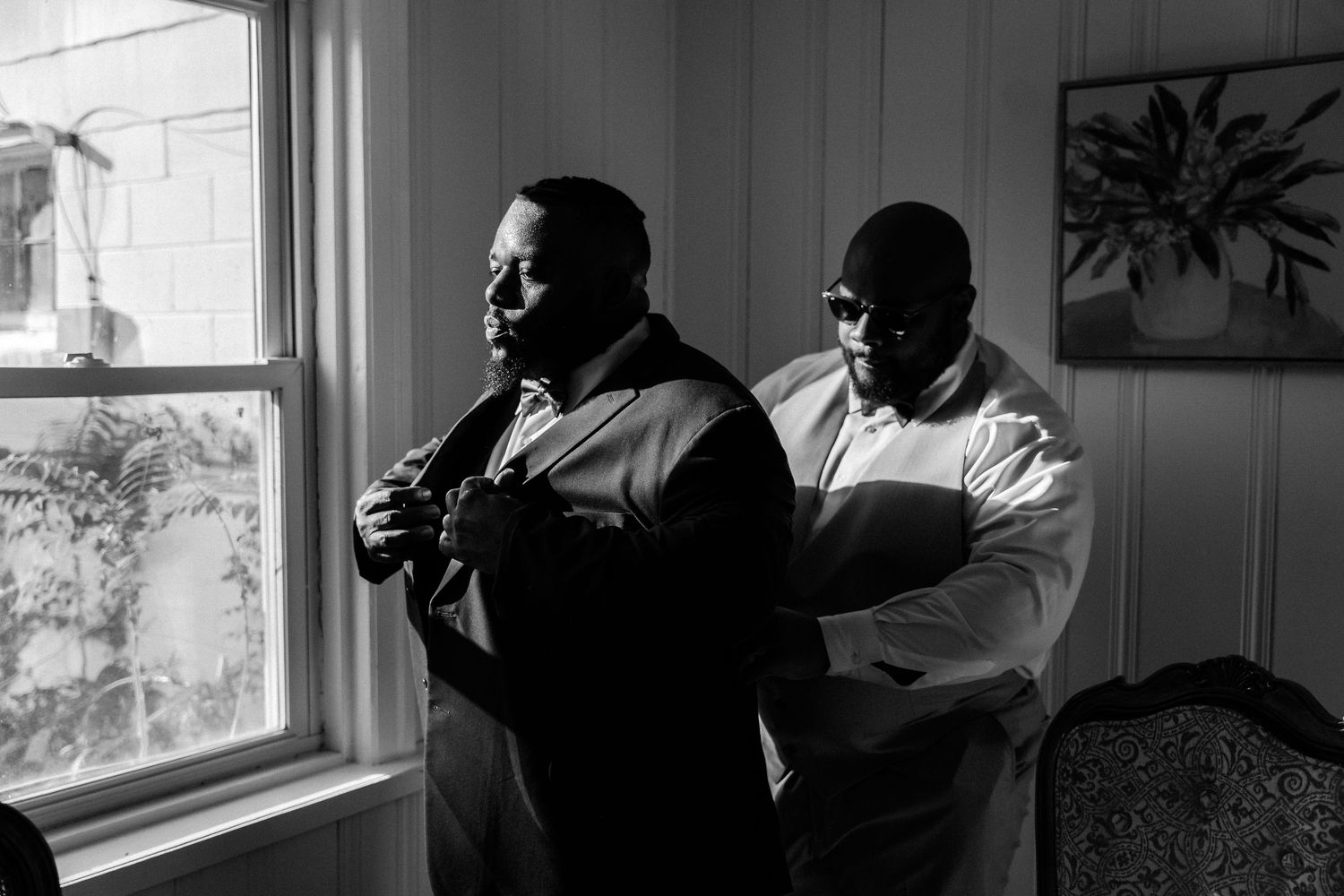 Two people in formal attire helping with wedding preparations in a sunlit room.