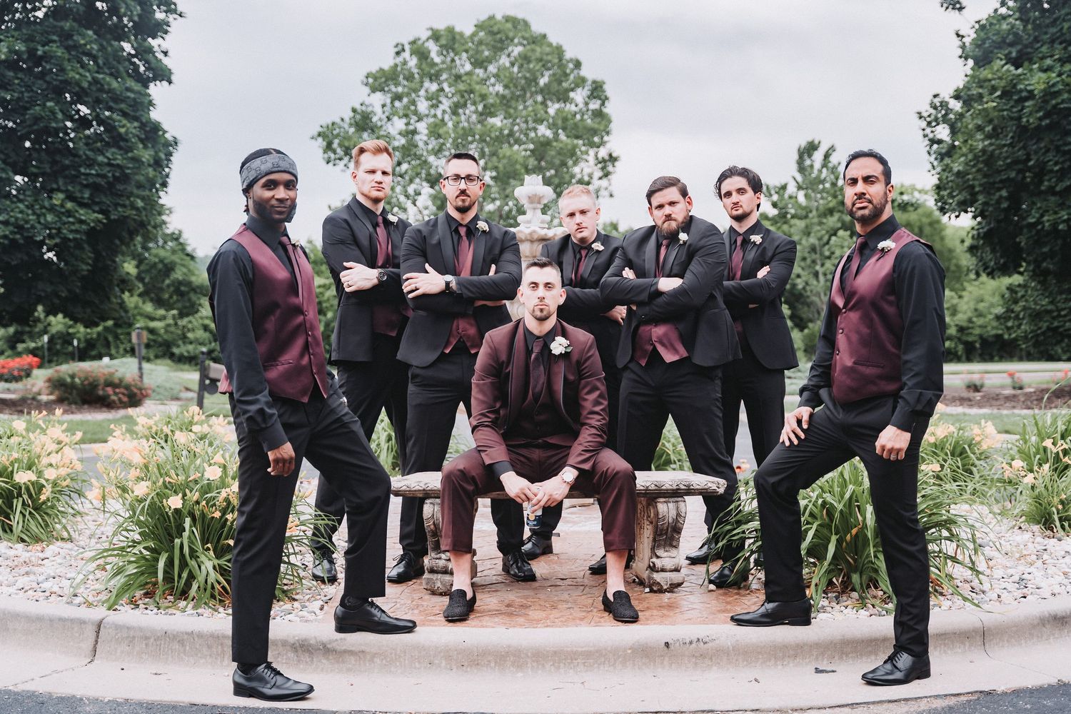 Wedding party in burgundy and black attire poses outdoors for formal group photo.