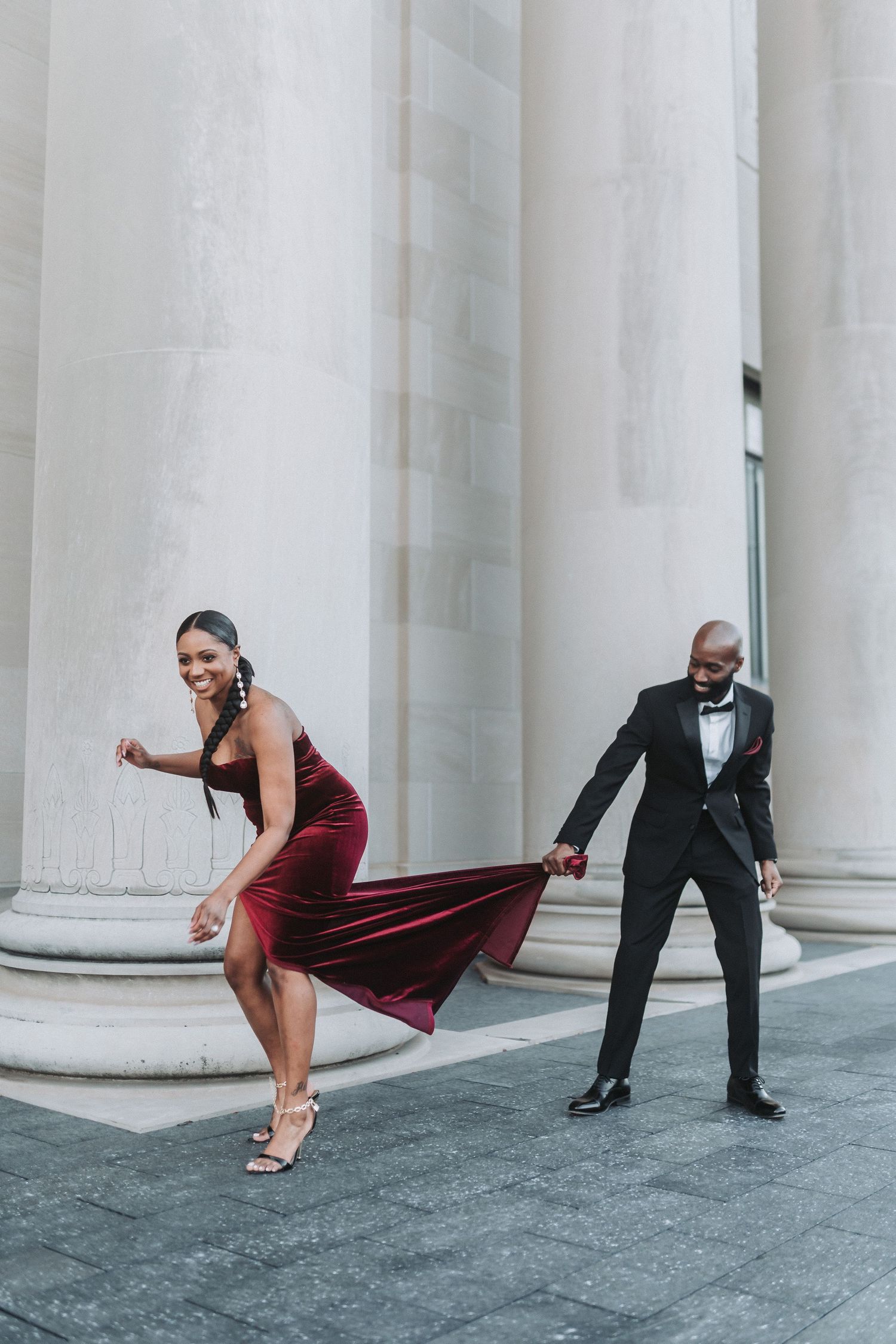 Playful couple in formal wear pose dramatically between white marble columns.