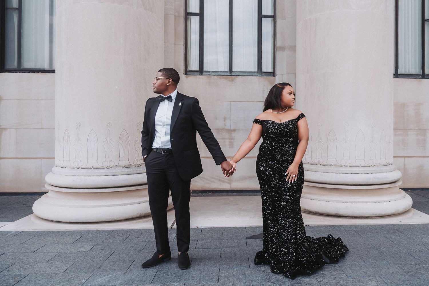 Stylish couple in black formal wear posing against architectural columns.
